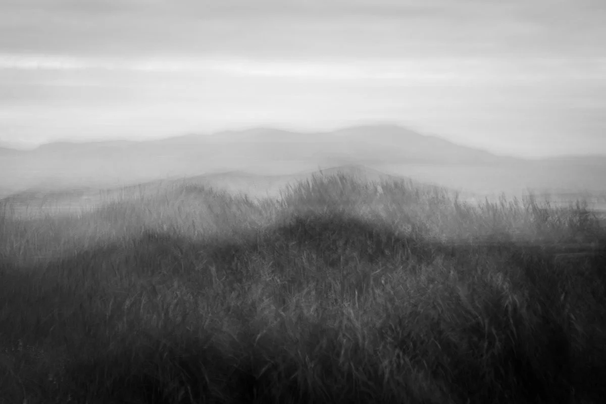 Black and white landscape photo of rolling hills and distant mountains with a misty or foggy appearance.