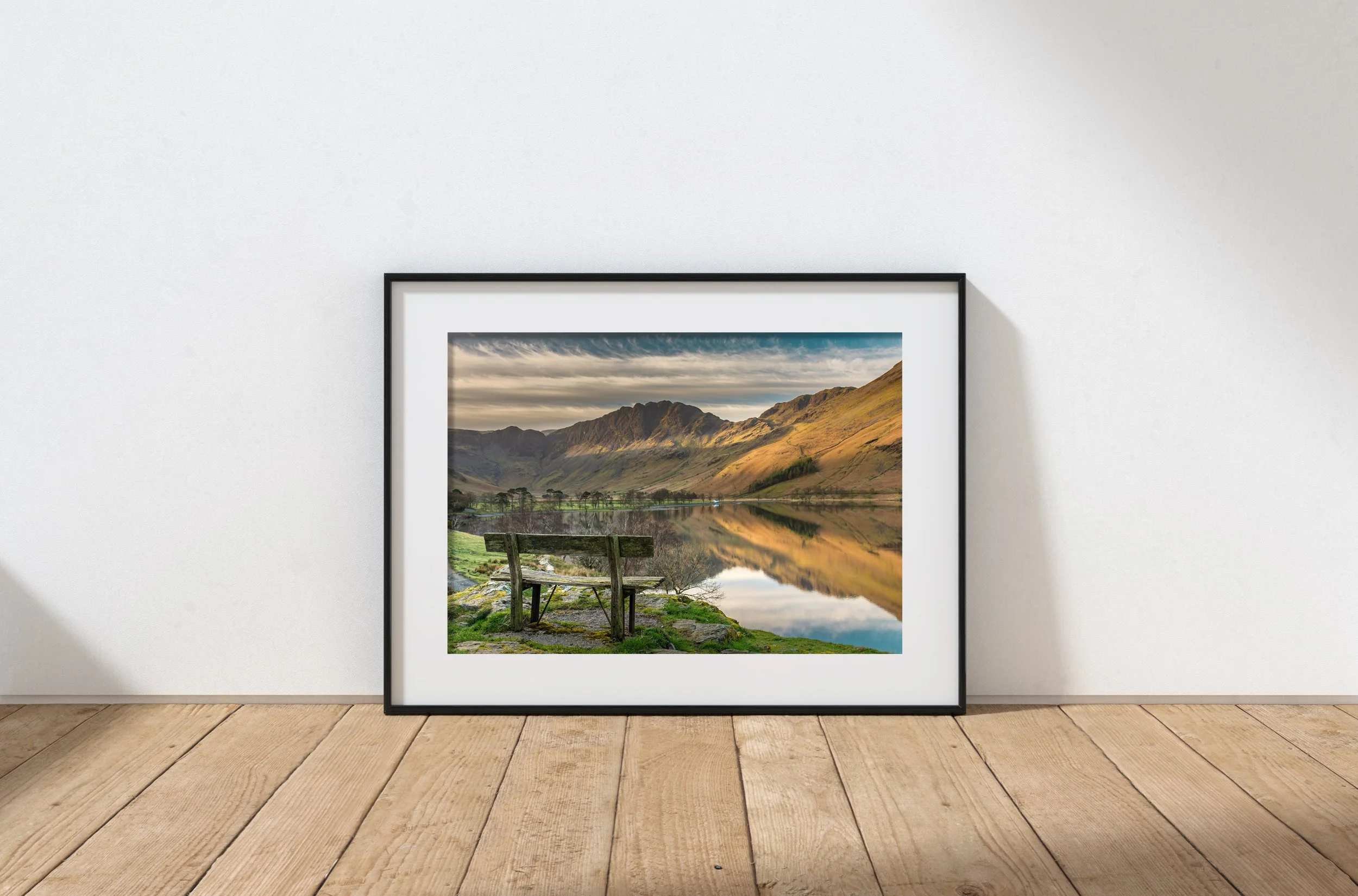 Framed landscape photograph of an old wooden bench overlooking Buttermere lake with mountains and a cloudy sky, leaning against a white wall on a wooden floor.