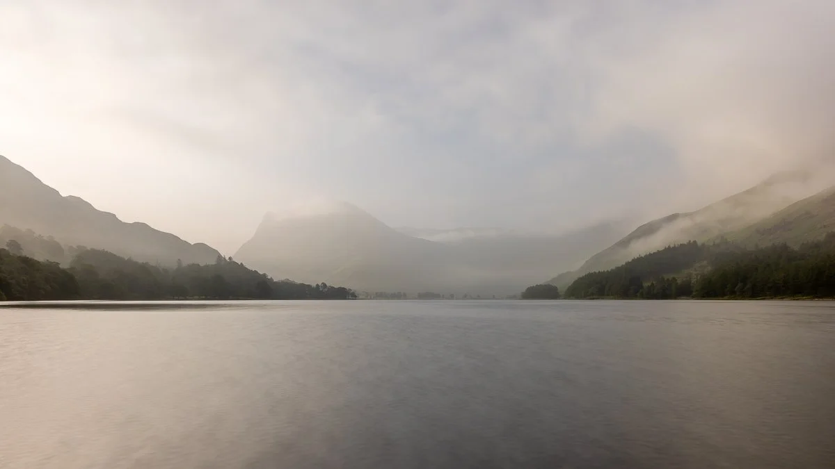 A long exposure smoothing Buttermere lake with misty green mountains under cloudy sky.