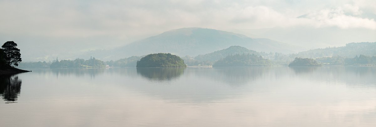 Calm lake with small islands, surrounded by misty mountains and cloudy sky.