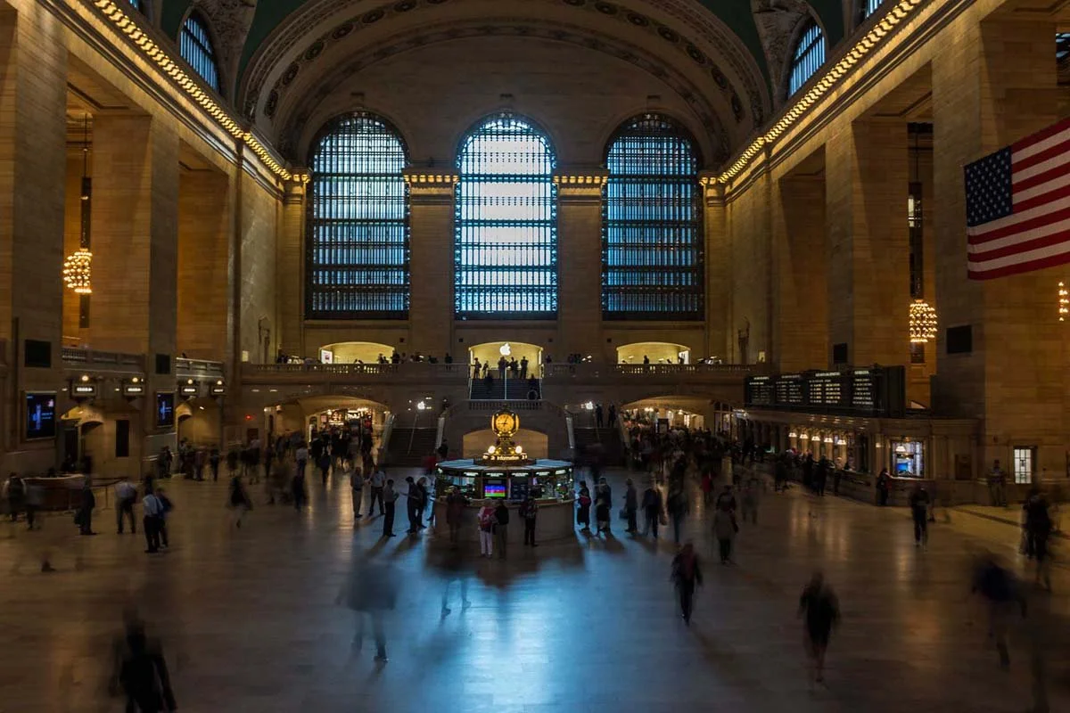 Inside Grand Central Train Station New York with high arched windows, people walking, and an American flag hanging on the wall.