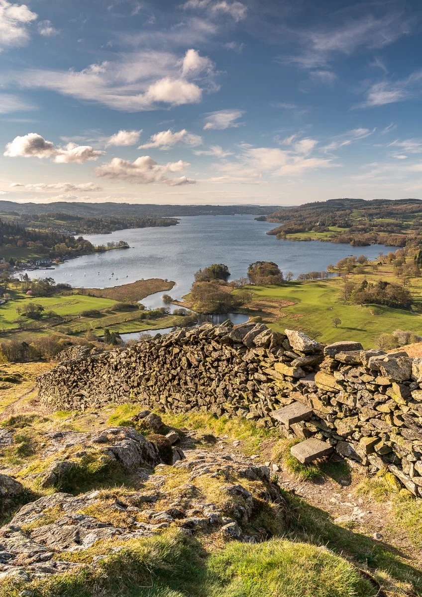 Scenic view of a lake surrounded by rolling green hills and farmland, with a stone wall in the foreground and partly cloudy sky taken from Todd Crag near Ambleside .