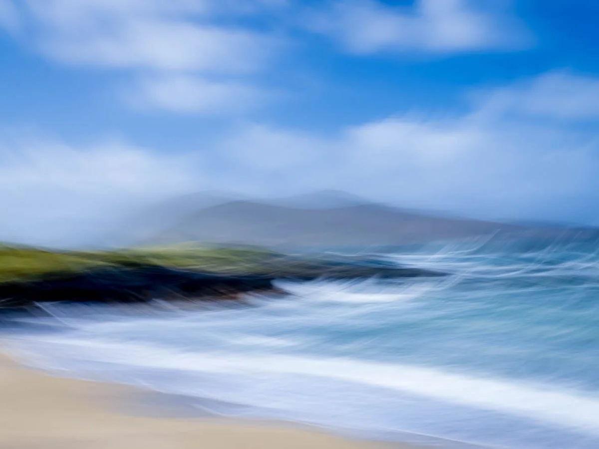 Blurry image of a coastal landscape with blue sky, clouds, green hills, and ocean waves.