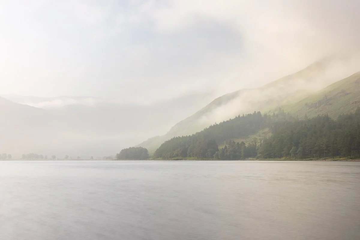 A foggy landscape with a large body of water in the foreground, lush green hills and mountains in the background, partially obscured by mist and clouds.