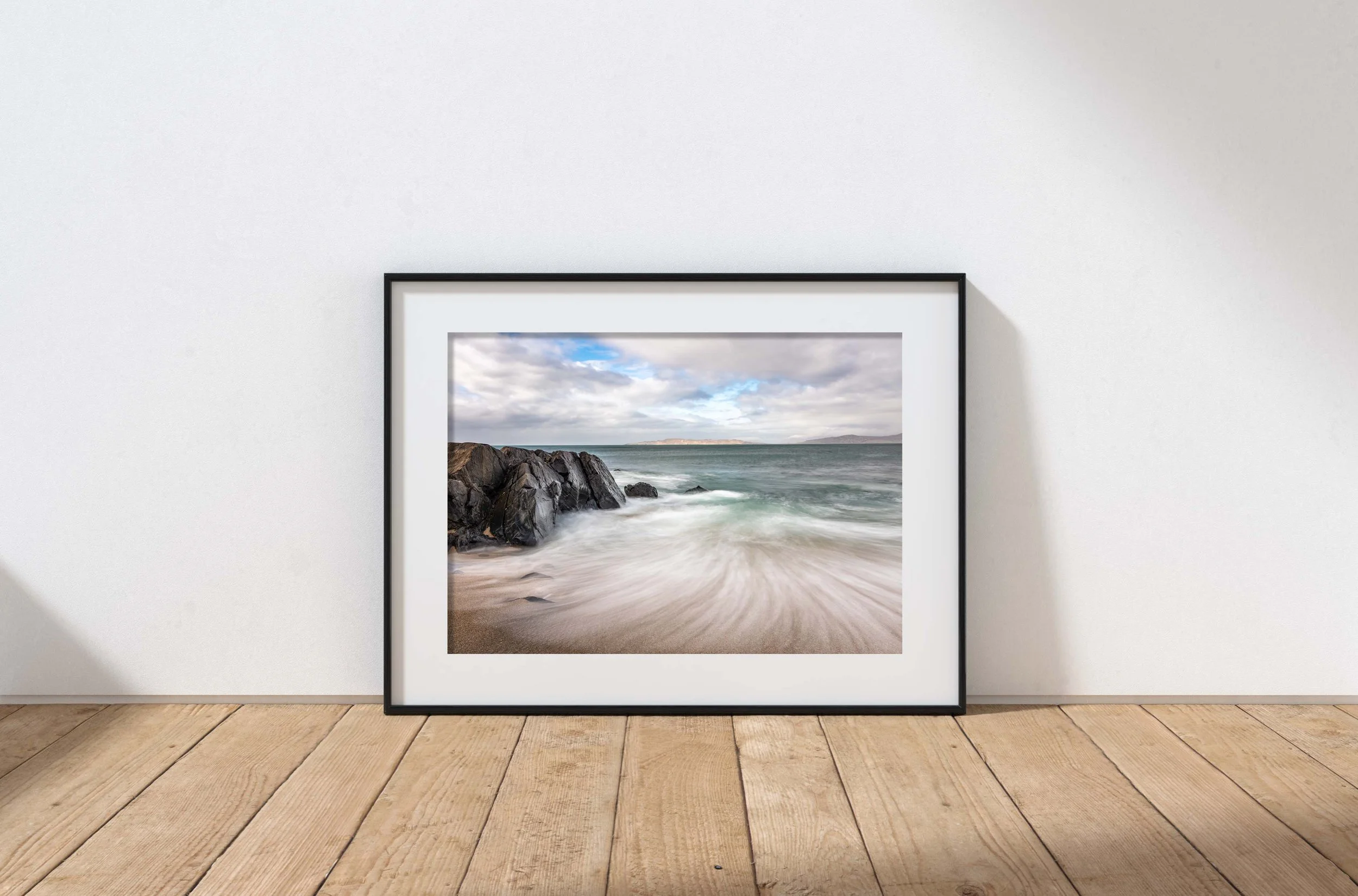 Framed ocean shoreline photograph resting on a wooden floor against a white wall.