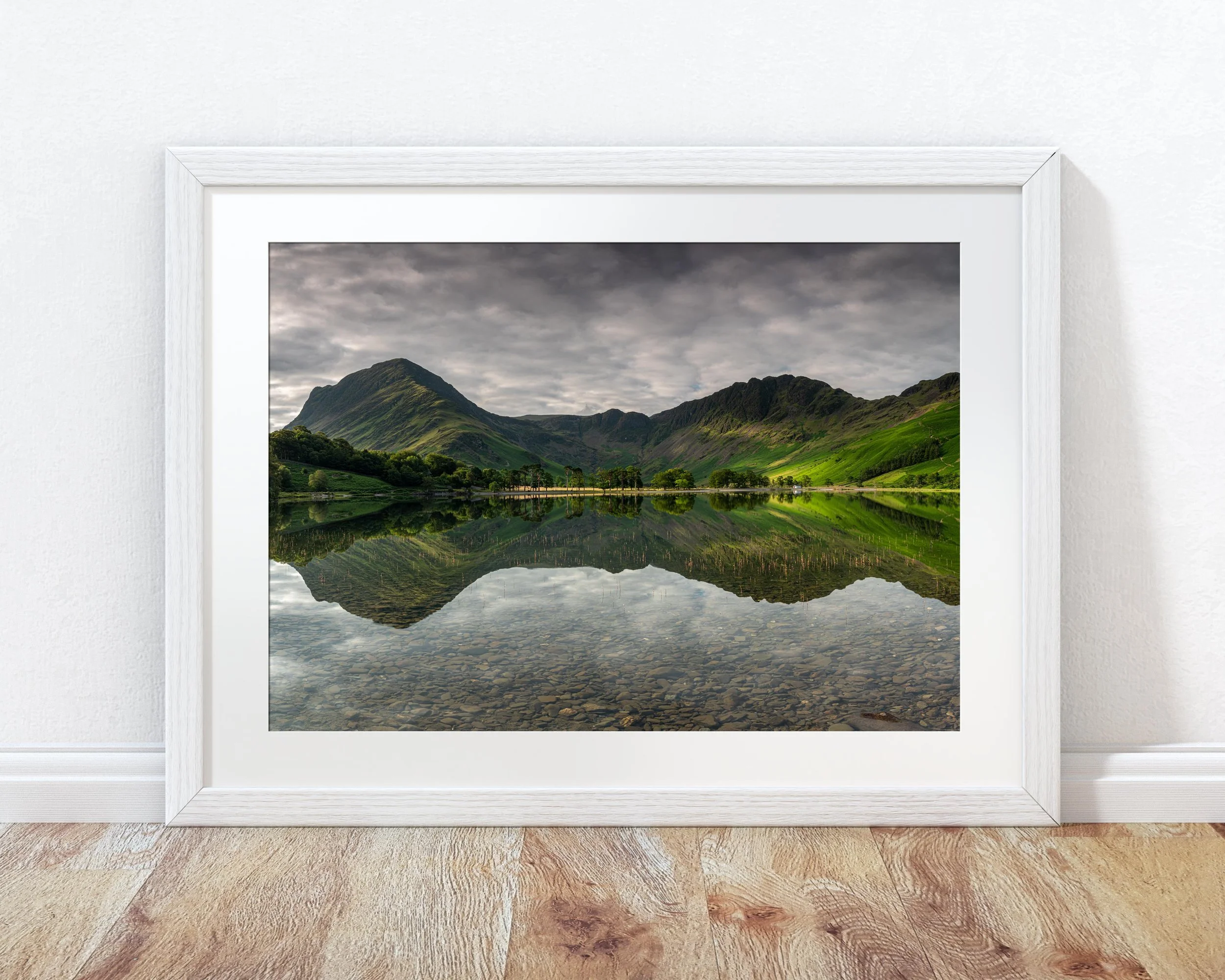 Framed landscape photograph of a mountain reflected on Buttermere lake with green hills and a cloudy sky, leaning against a white wall on wooden flooring.