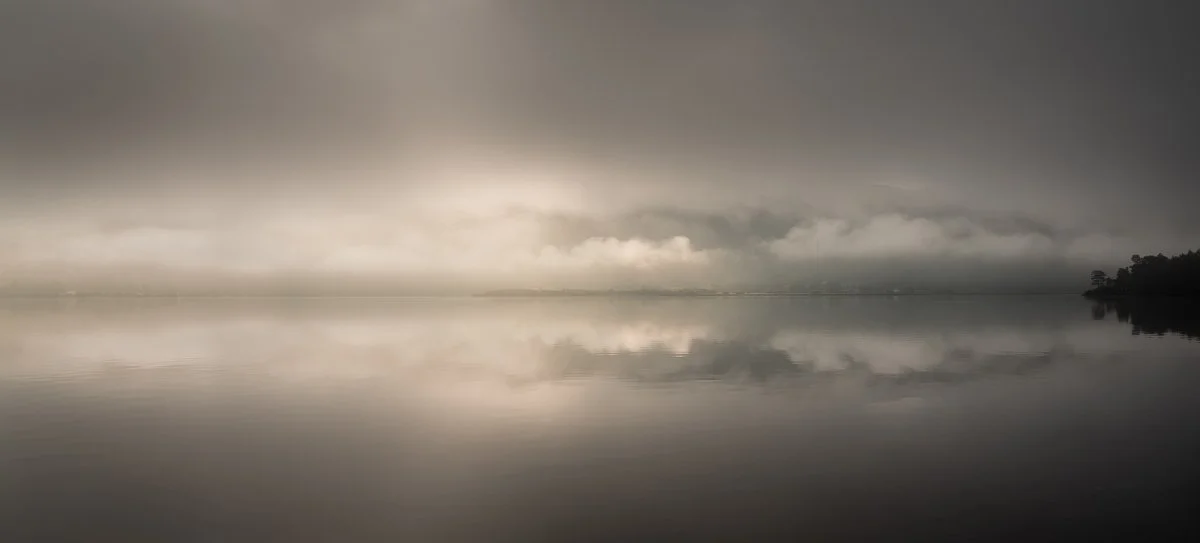 A calm, reflective body of water under an overcast sky with dark clouds and a faint horizon line in the distance.