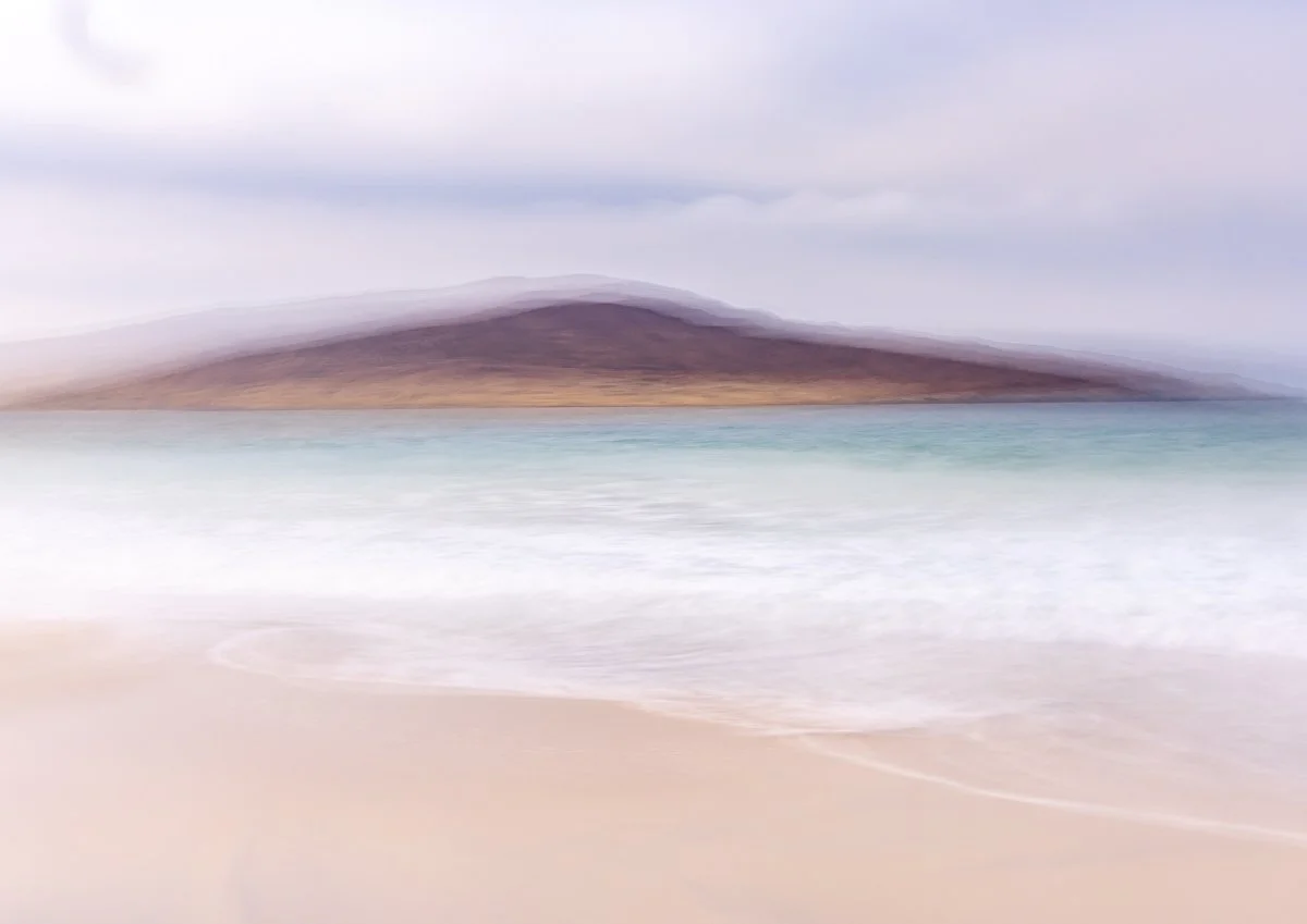 A beach scene with a sandy shoreline, ocean waves, and a distant landmass or mountain under an overcast sky.