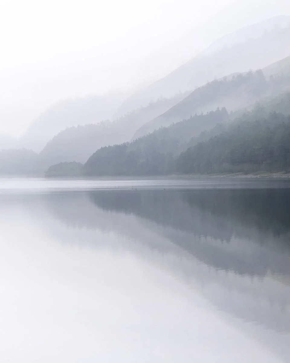 A misty landscape featuring calm water with a perfect reflection, surrounded by rolling, forested hills and mountains in the background.