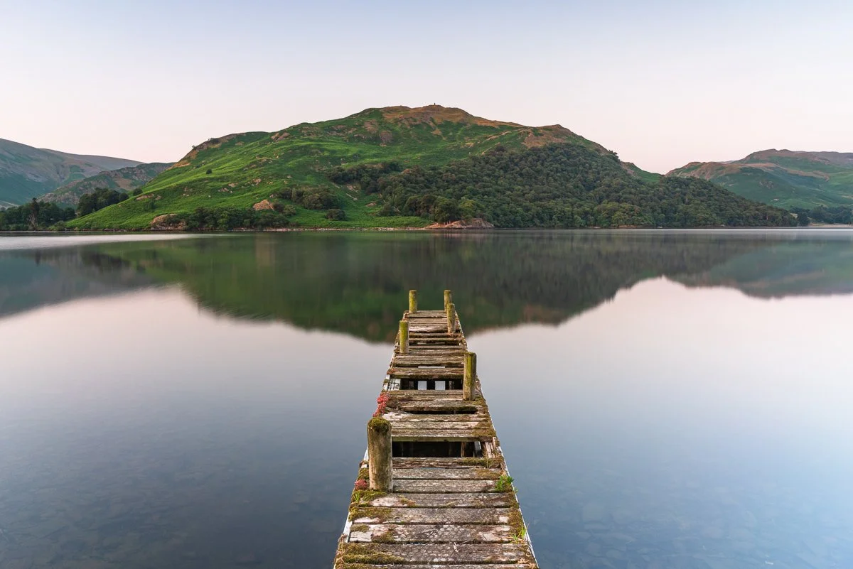 An old wooden dock on Ullswater extending into a calm lake, with green hills and a mountain in the background, under a clear sky.