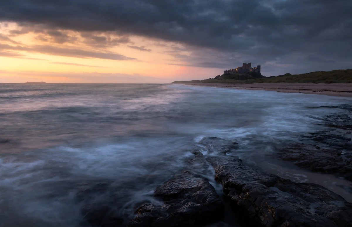 A coastal scene at sunset with waves hitting rocks in the foreground, dark clouds overhead, and a castle on a hill in the distance.