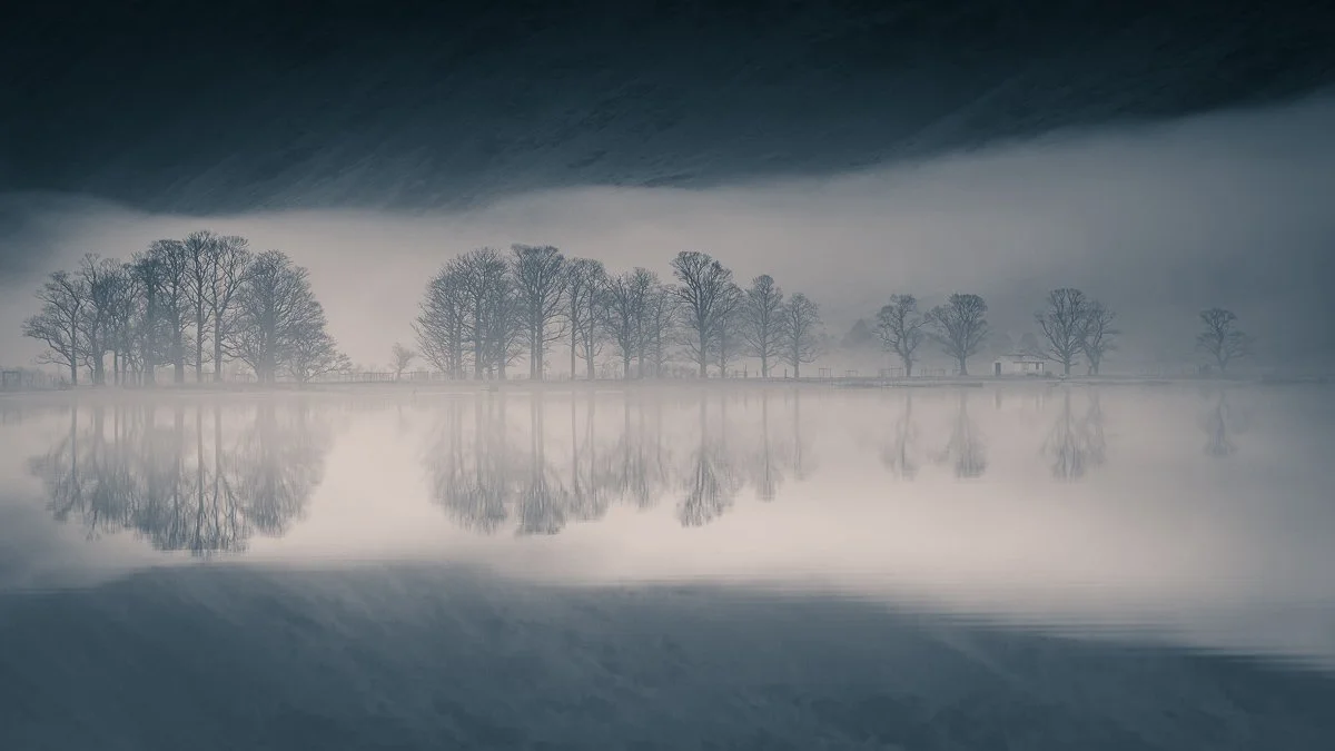 A misty scene with trees reflected in Buttermere lake.