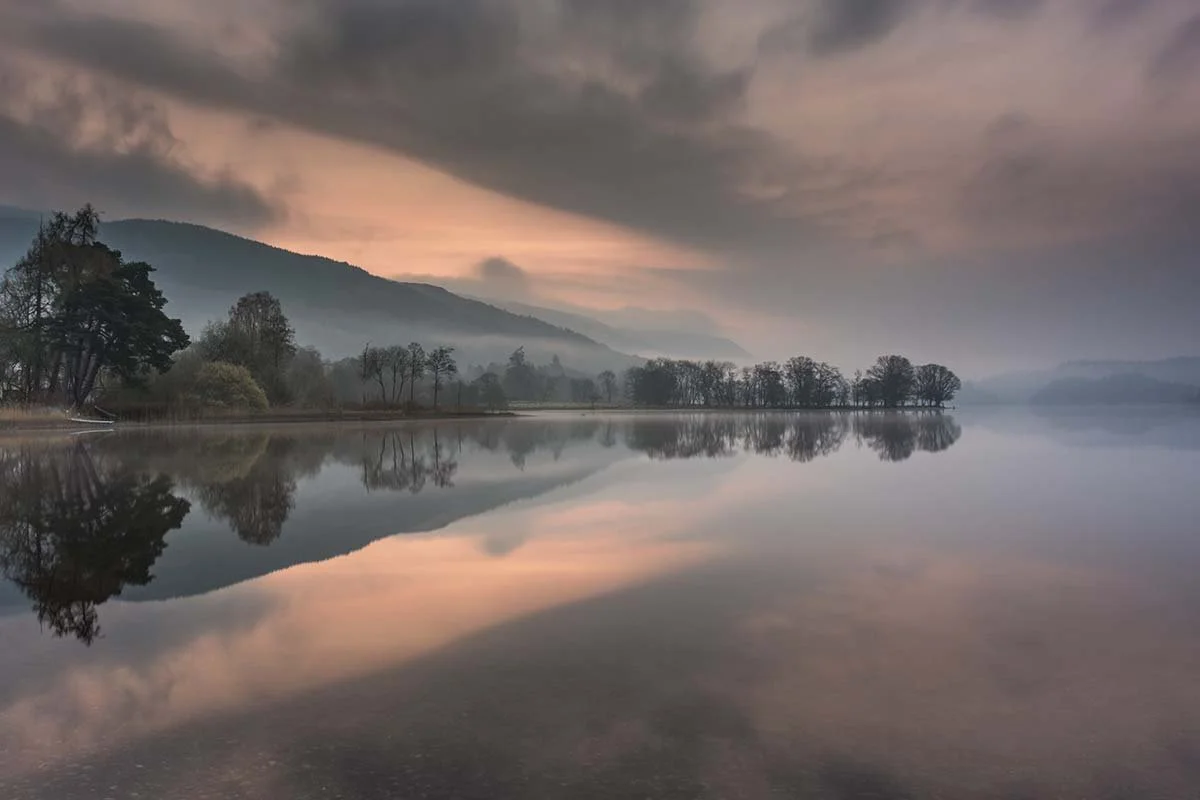 A misty scene on Loch Ard in the Trossachs with calm water and reflections and muted peach colours in the sky and water.