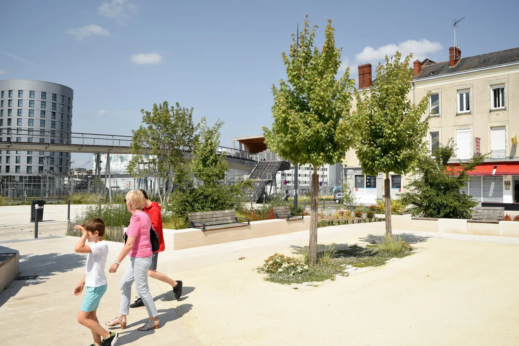 Une placette urbaine avec des arbres, des bancs en bois, et des enfants qui jouent ou se promènent par une journée ensoleillée PRISME Photographie