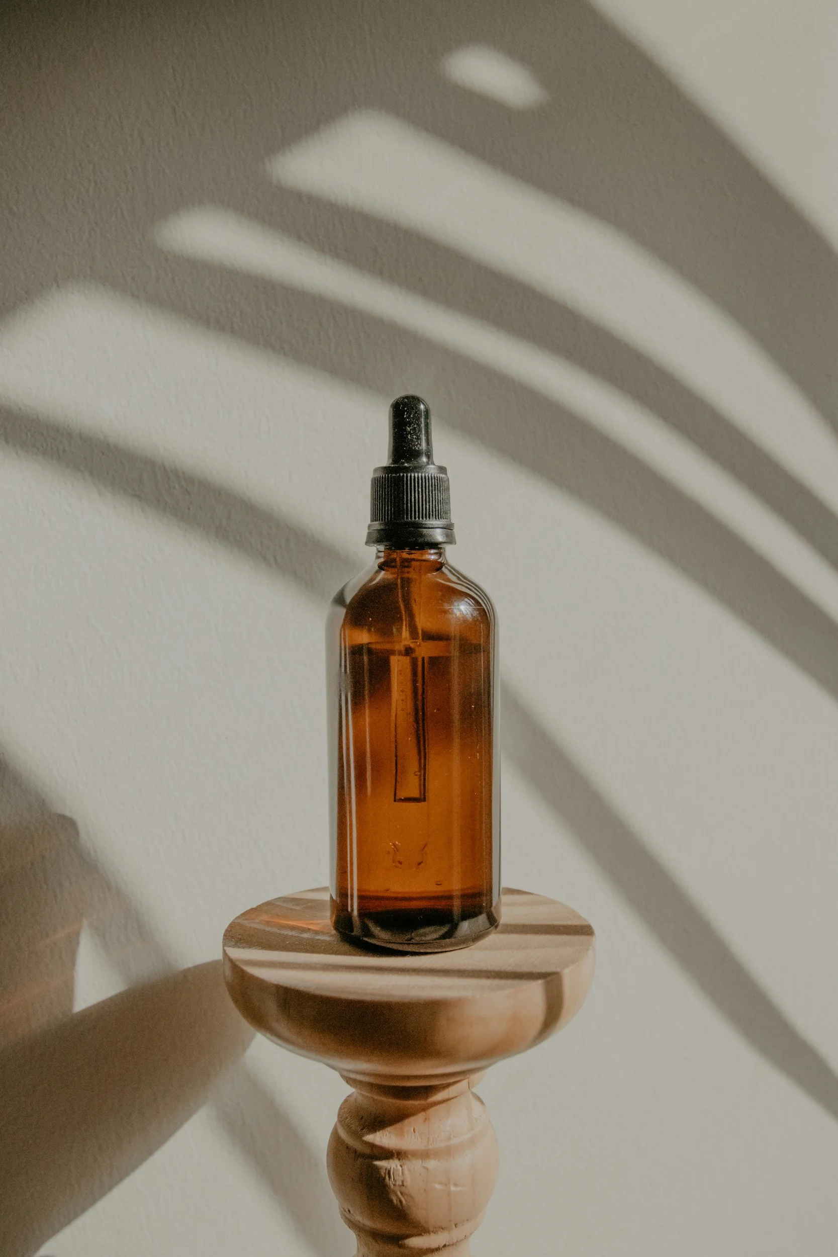An amber glass spray bottle with a black dropper cap on a wooden surface, casting a patterned shadow on a light-colored wall.
