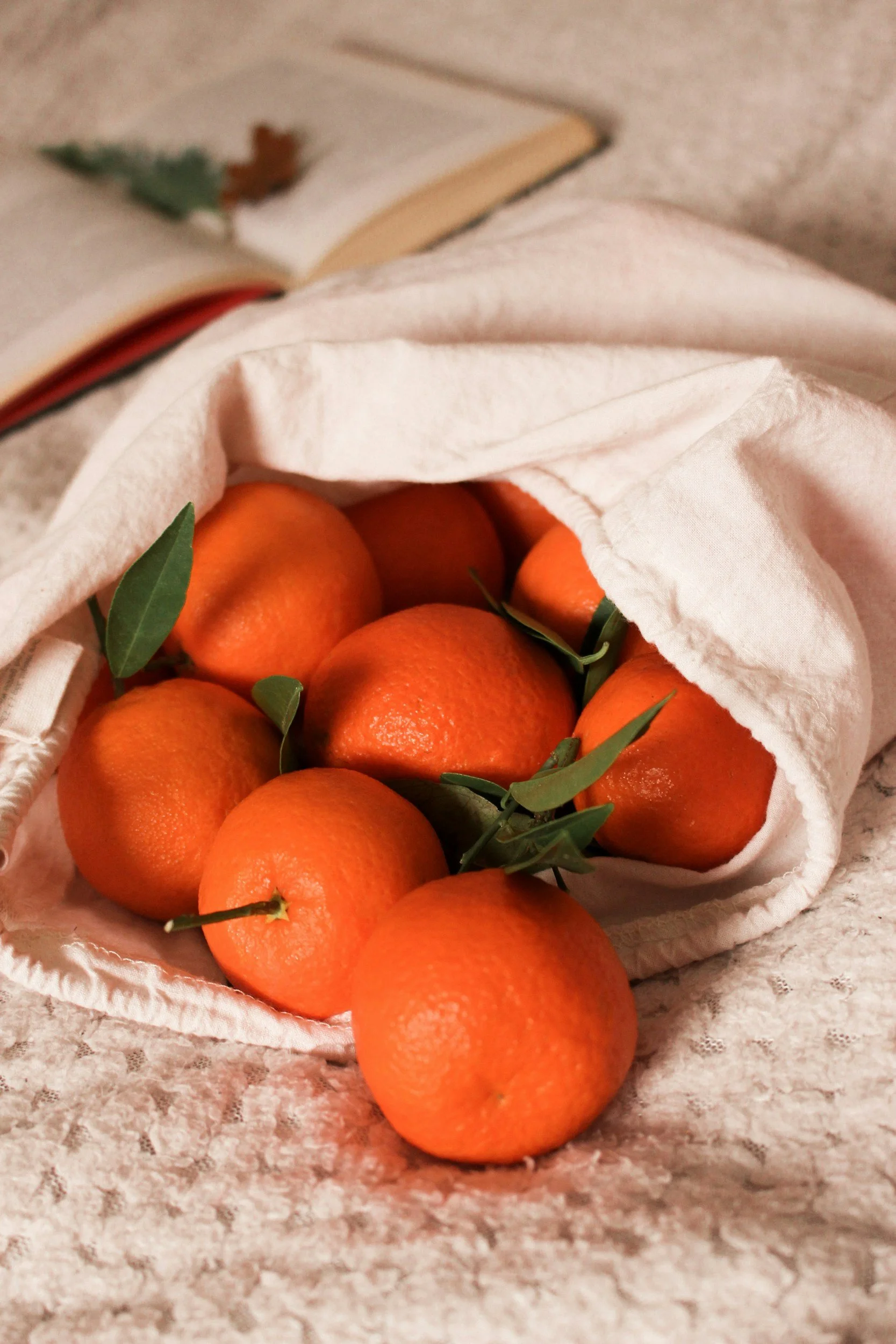 A white cloth bag filled with fresh, ripe oranges, some with green leaves still attached, resting on a lace surface.