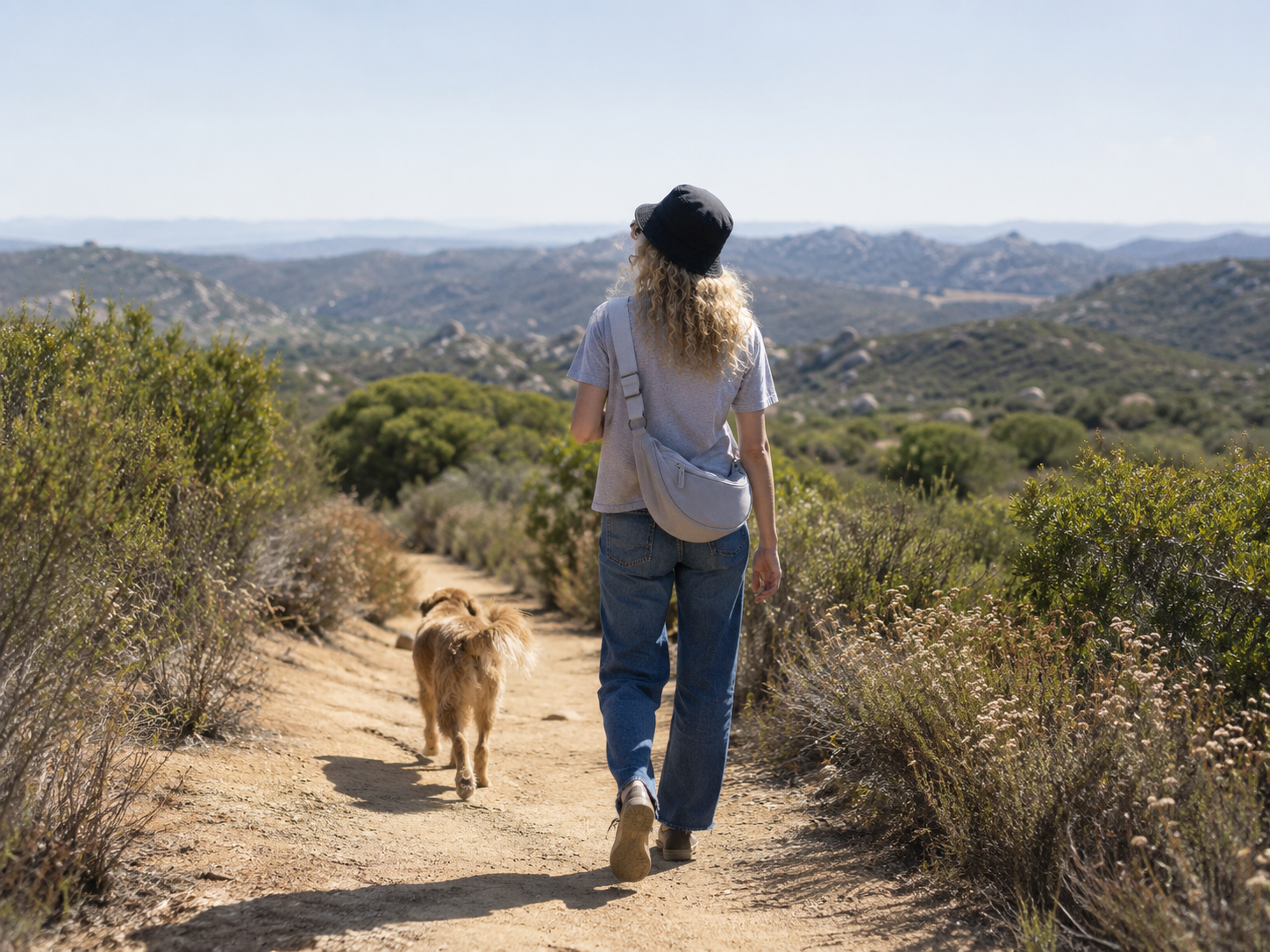A woman walking with a dog on a dirt trail through a desert landscape with mountains in the background.