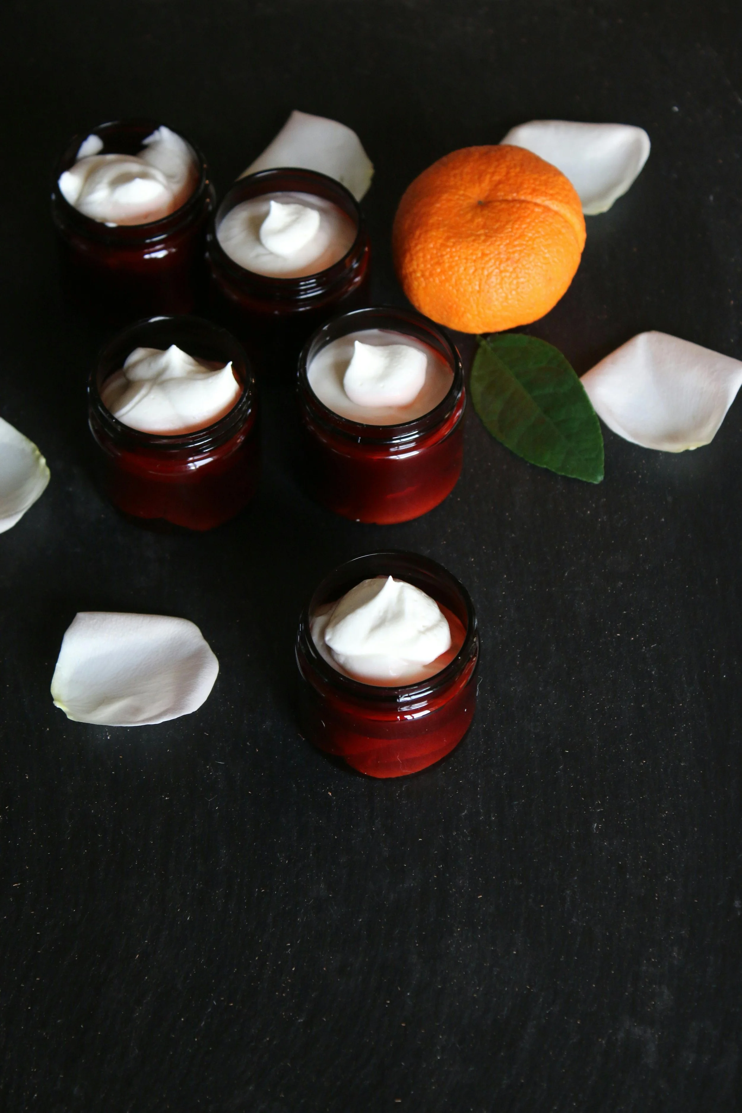 Four small jars of white cream, a whole orange with a green leaf and white flower petals surrounding it on a black surface.