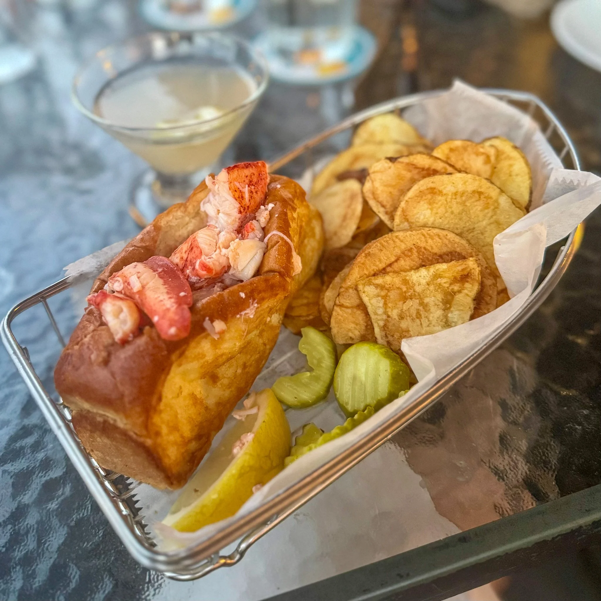 Lobster roll with side of potato chips and pickle slices on a metal tray, with a martini glass in the background.