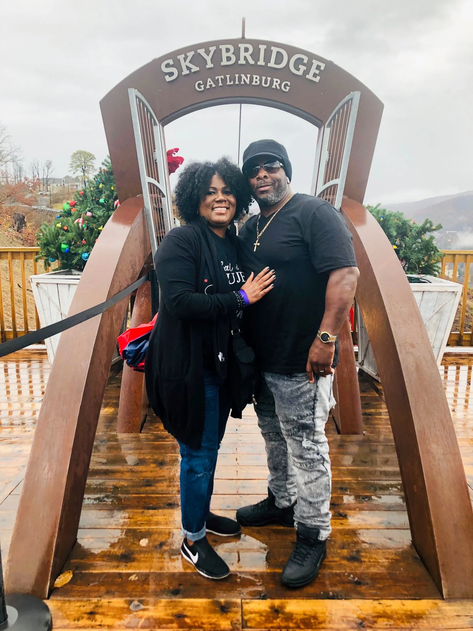 A smiling couple posing together on the SkyBridge at Gatlinburg during overcast weather, with Christmas trees and mountains in the background.