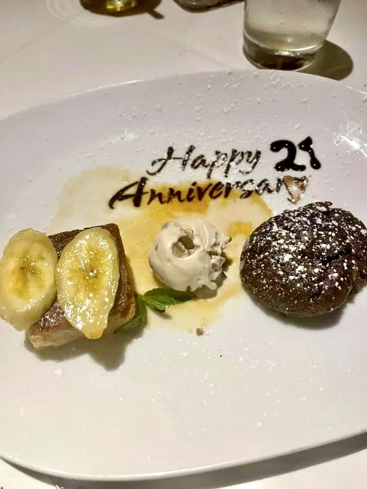 Dessert plate with banana slices, a small cake or pastry, a dollop of whipped cream, and a chocolate cookie, with a message reading 'Happy 25th Anniversary' written in chocolate.