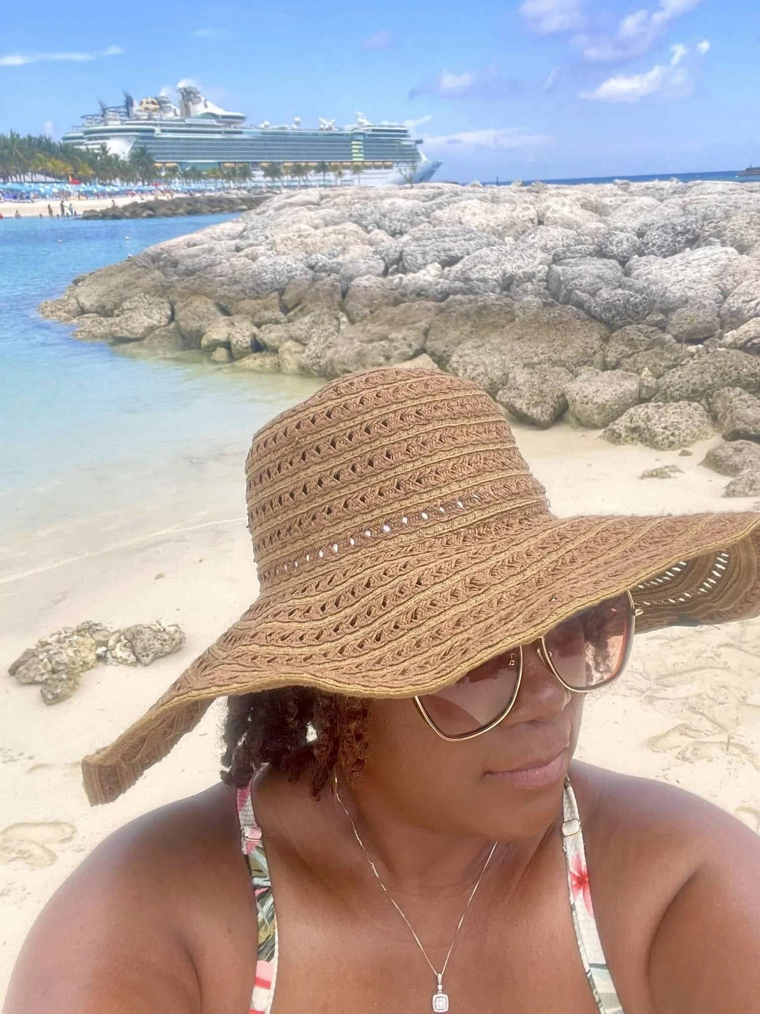 Woman wearing a large sunhat and sunglasses on a beach with rocks, ocean, and cruise ship in the background.