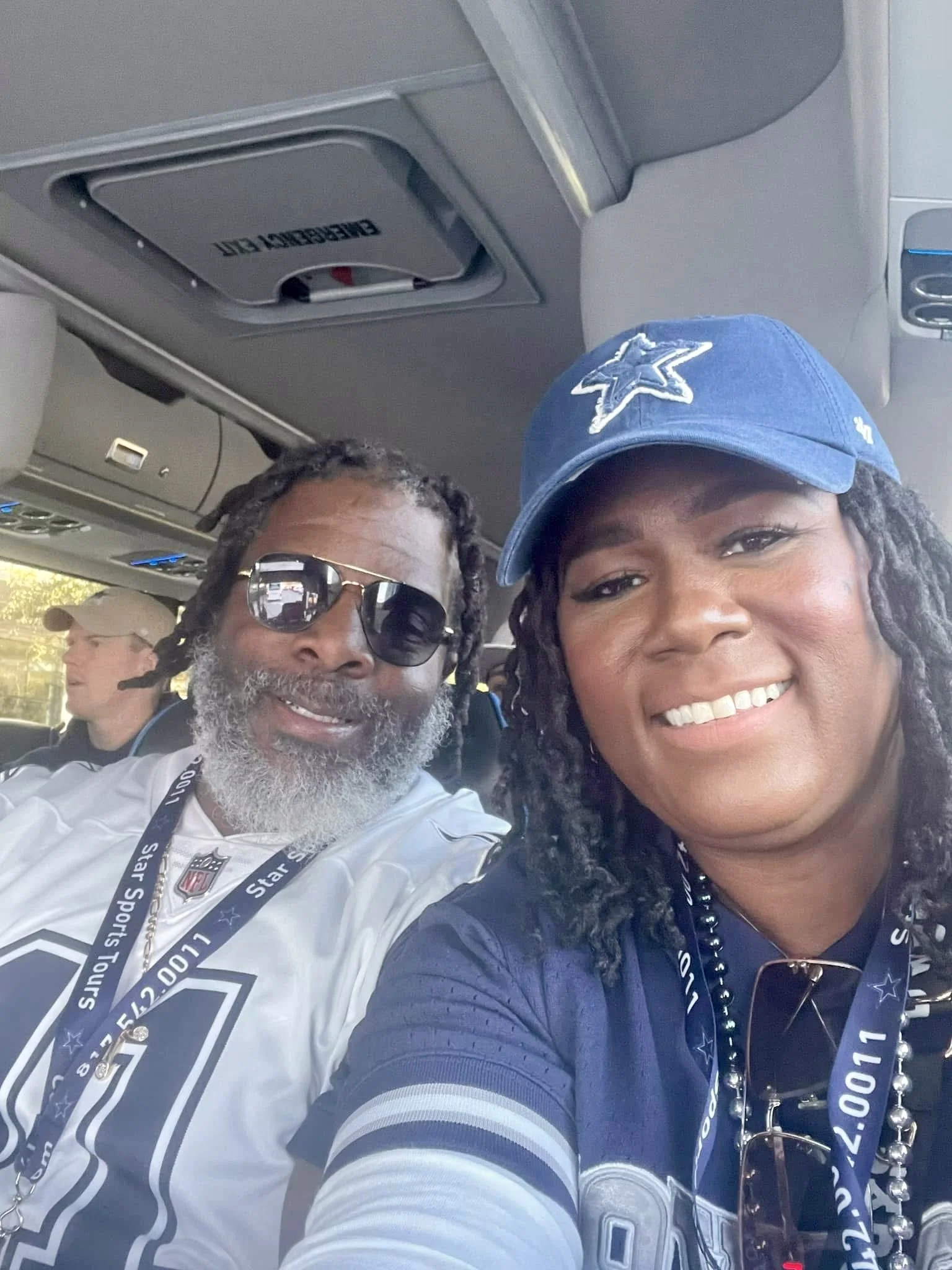 A smiling woman wearing a Dallas Cowboys cap and a woman with sunglasses and a Dallas Cowboys shirt sitting inside a bus, with another person faintly visible in the background.