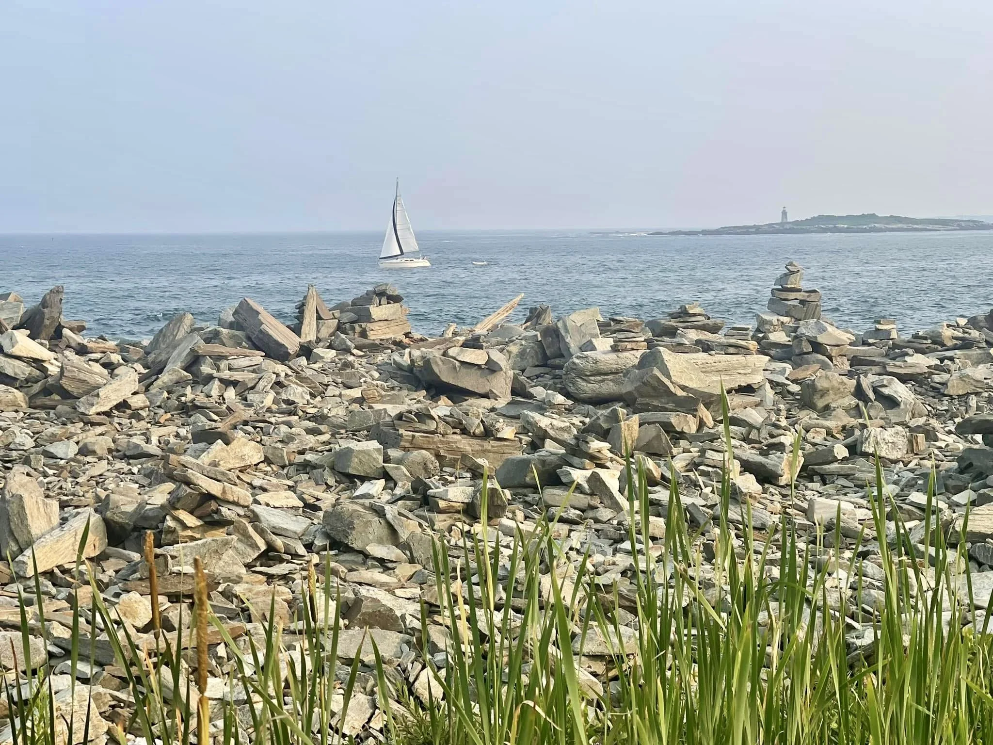 Rocky shoreline with green grass in foreground and a sailboat in the ocean, with a lighthouse on a distant island.