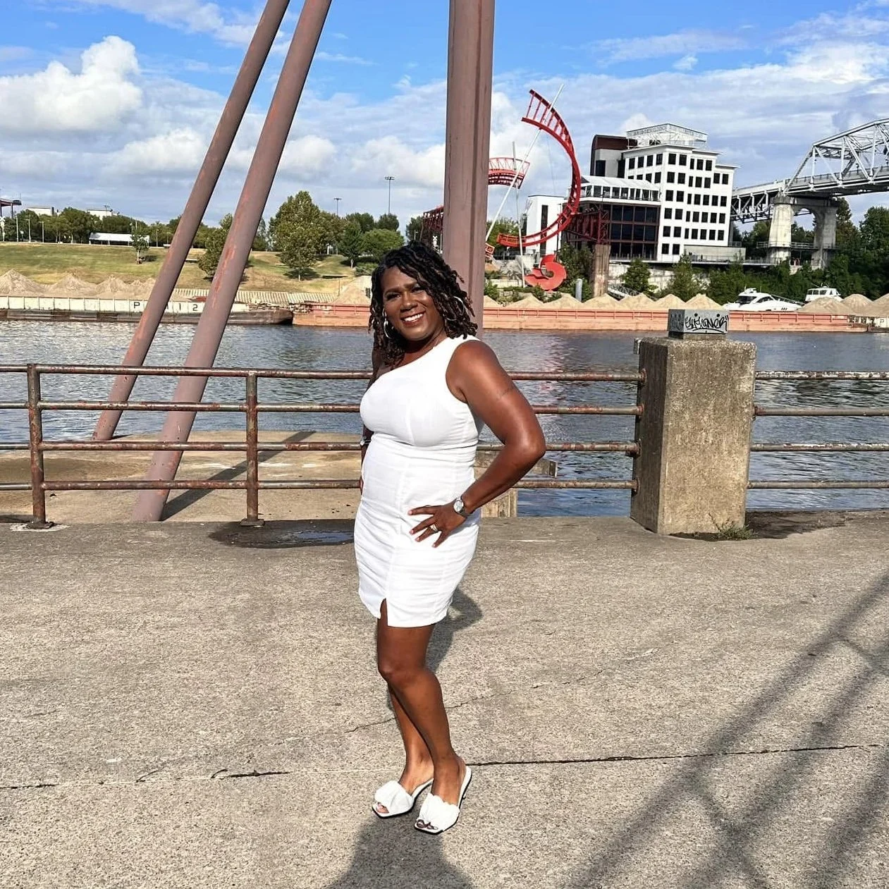 A woman in a white dress and high heels standing on a concrete path near a body of water, with modern buildings and a large red sculpture in the background.