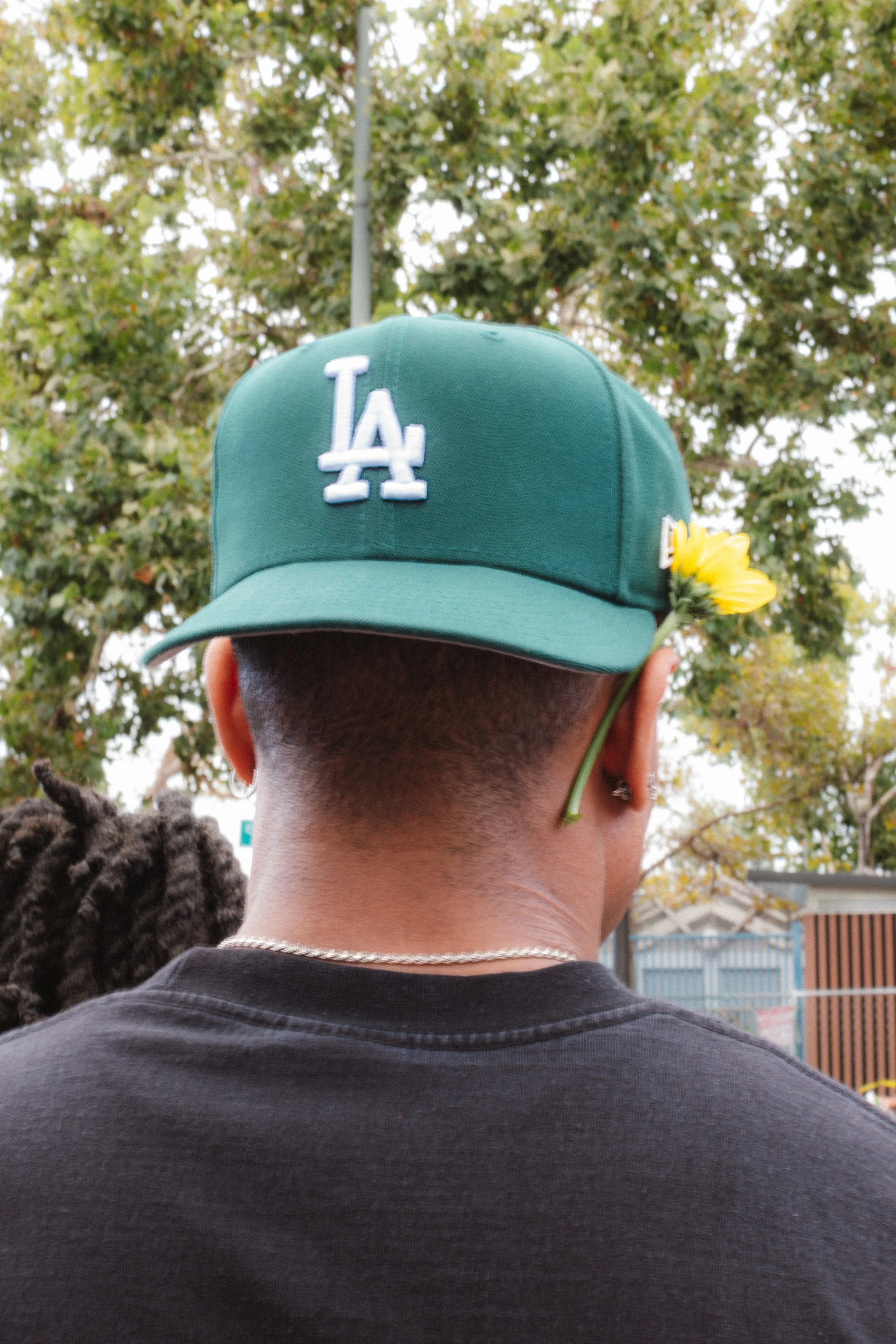 Back of a person's head wearing a green Los Angeles Dodgers baseball cap, with a yellow flower tucked into the cap's strap, outdoors with trees in the background.