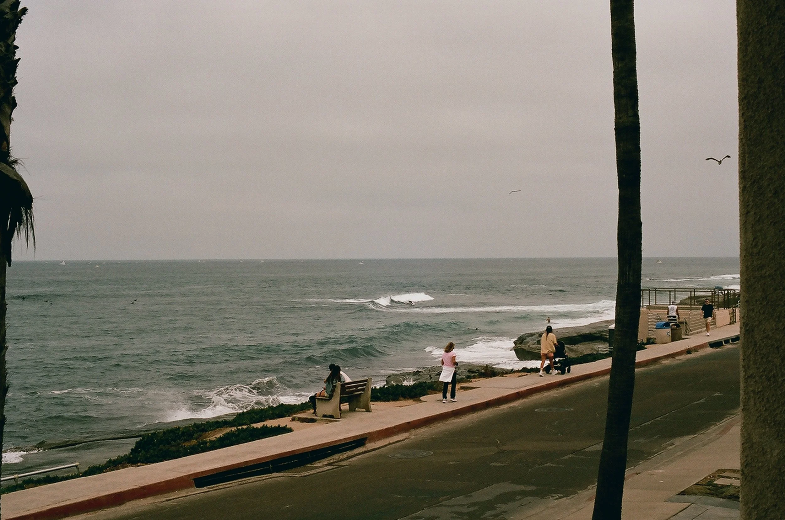 La Jolla overcast day at the beach with people walking along the sidewalk, some sitting on benches near the rocky shoreline, and ocean waves with a few surfers.