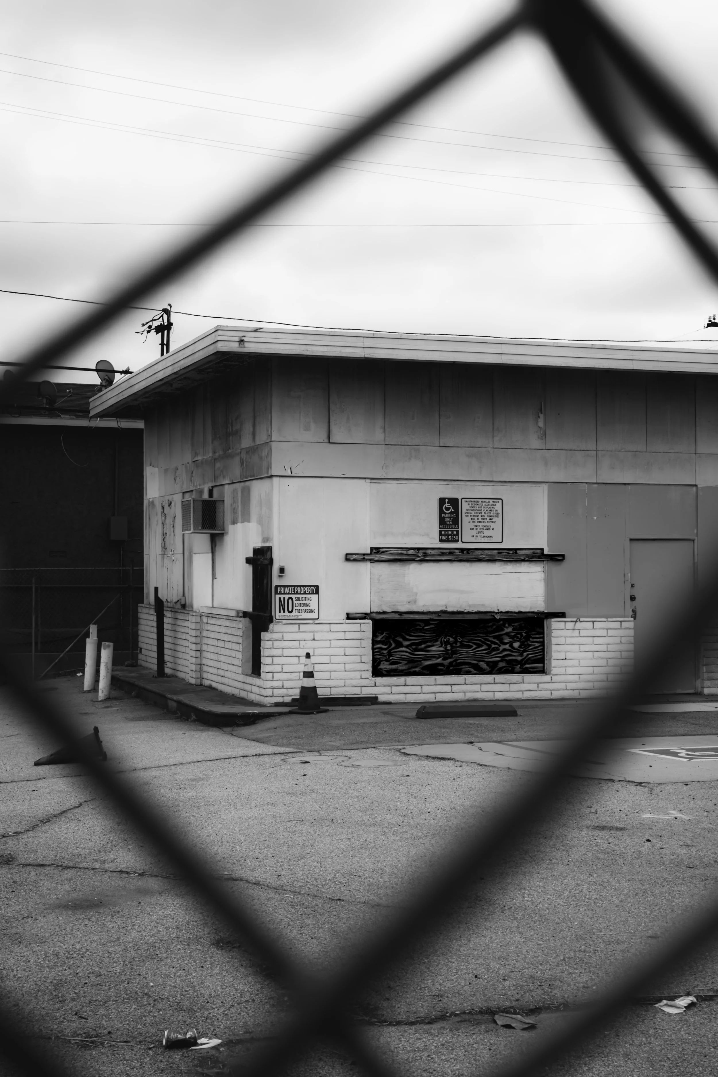 Black and white photo taken through a chain-link fence, showing a small, abandoned building with boarded-up windows and a sign indicating parking restrictions. The sky is overcast, and the ground is cracked with some litter.