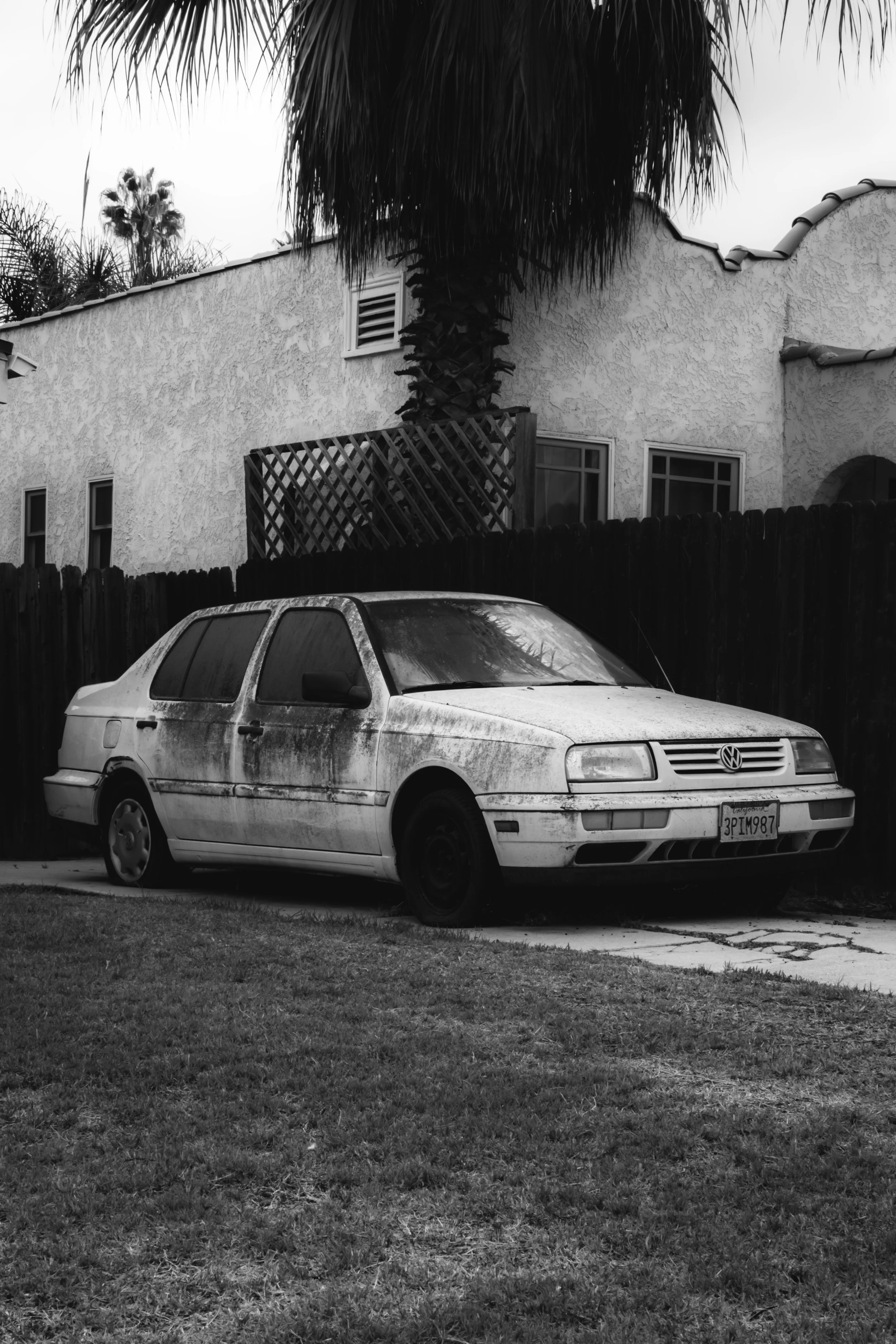 A black-and-white photograph of an old, dirty Volkswagen hatchback car parked in a yard with a house and a tall palm tree in the background. The car is covered in grime and has a worn appearance.