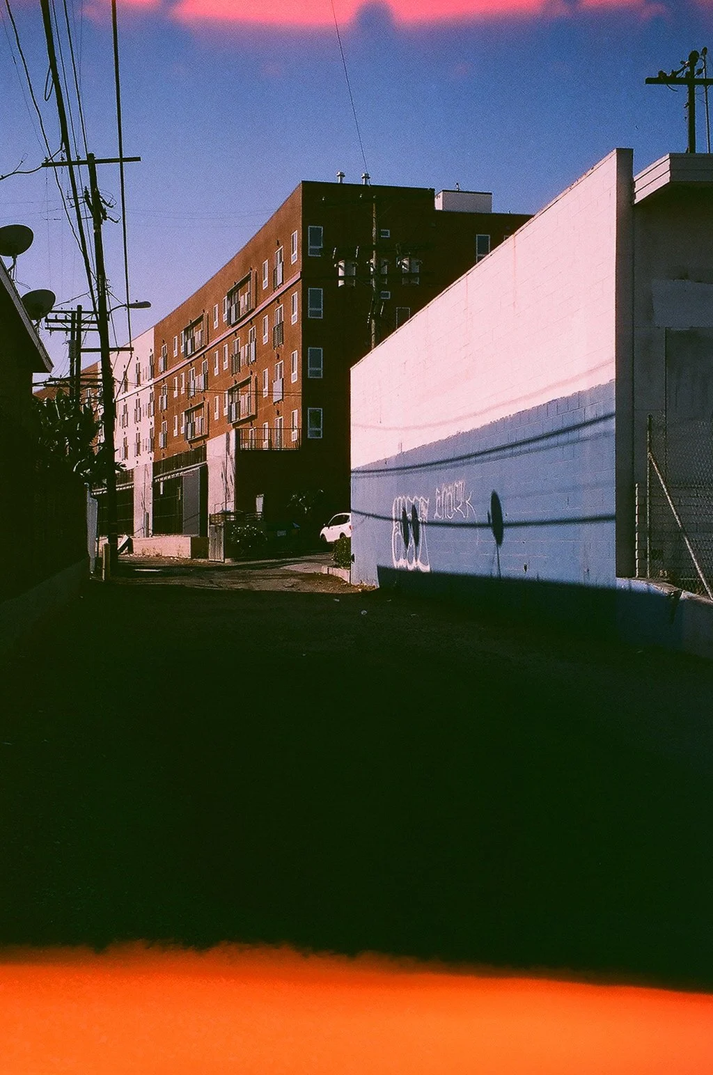 An alleyway during sunset with a multi-story brick building on one side and a white wall with graffiti on the other. Utility poles and power lines are visible against a sky with pink and orange hues.