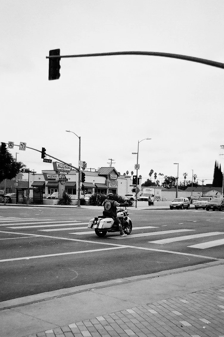 A person riding a motorcycle stopped at a crosswalk at an intersection, with traffic lights and commercial buildings in the background, in black and white.