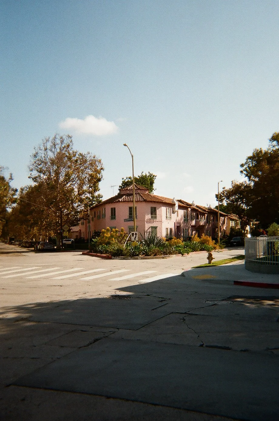 A residential neighborhood featuring a pink two-story building with a tiled roof, surrounded by trees and plants, under a clear blue sky with a few clouds.