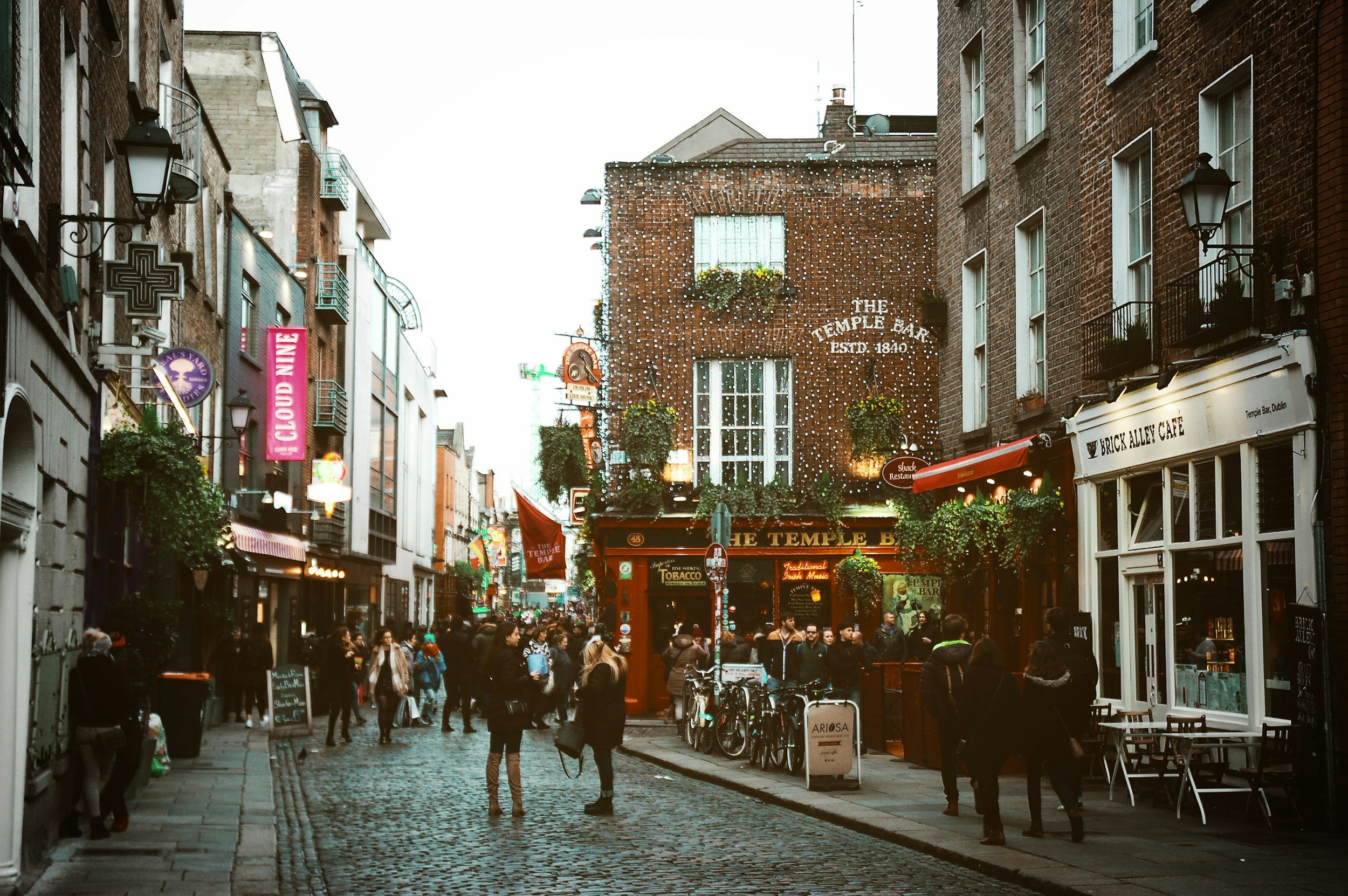 A busy cobblestone street in a city, lined with various bars, cafes, and shops with signs and hanging plants. Crowds of people walk and gather outside establishments, with bicycles parked in front of one. The buildings are multi-story with brick and modern facades.