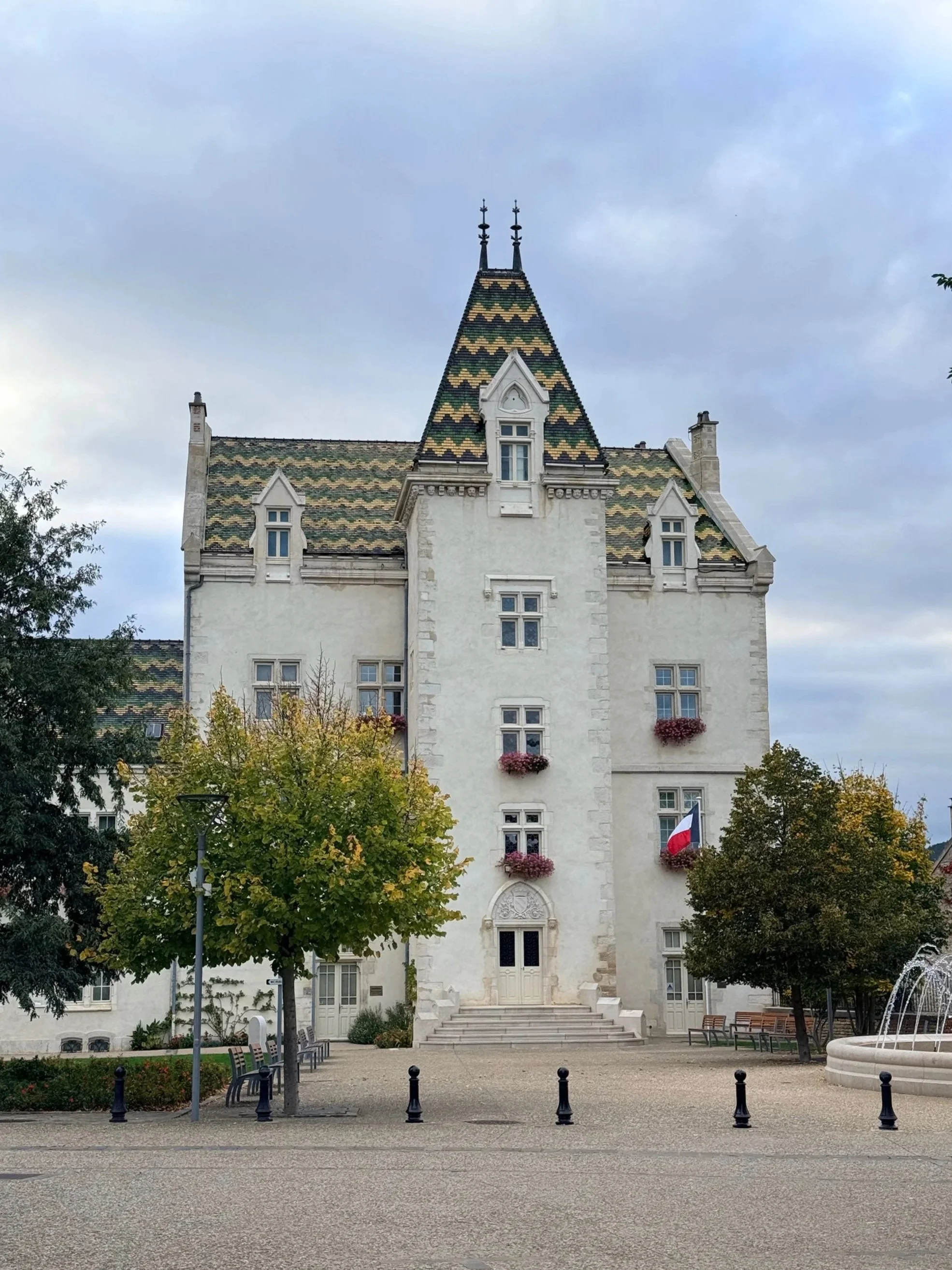 A historic white castle with a steep, colorful tiled roof and small windows, surrounded by trees, benches, and a fountain in a town square.
