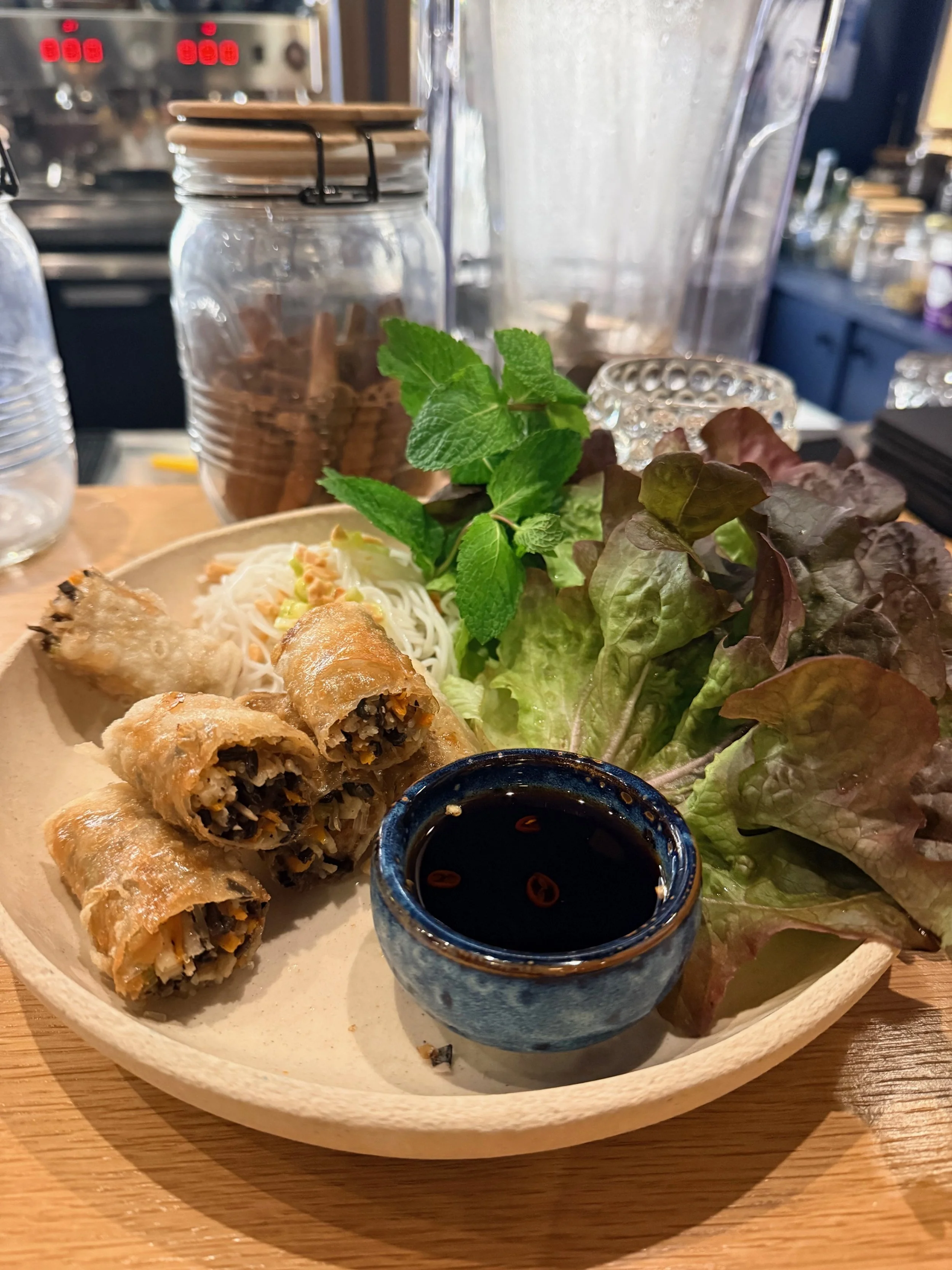 A plate of spring rolls with dipping sauce, served with mixed greens and fresh mint leaves on a wooden table.