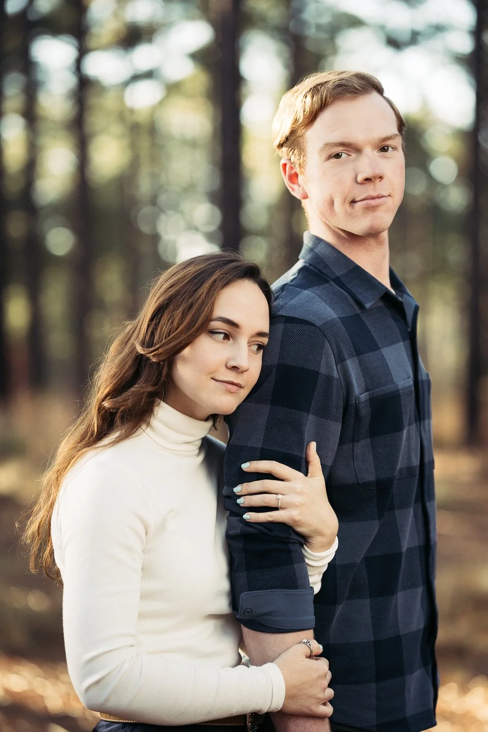 A young woman with long brown hair and light skin hugging a tall young man with red hair and light skin in a forest during autumn.