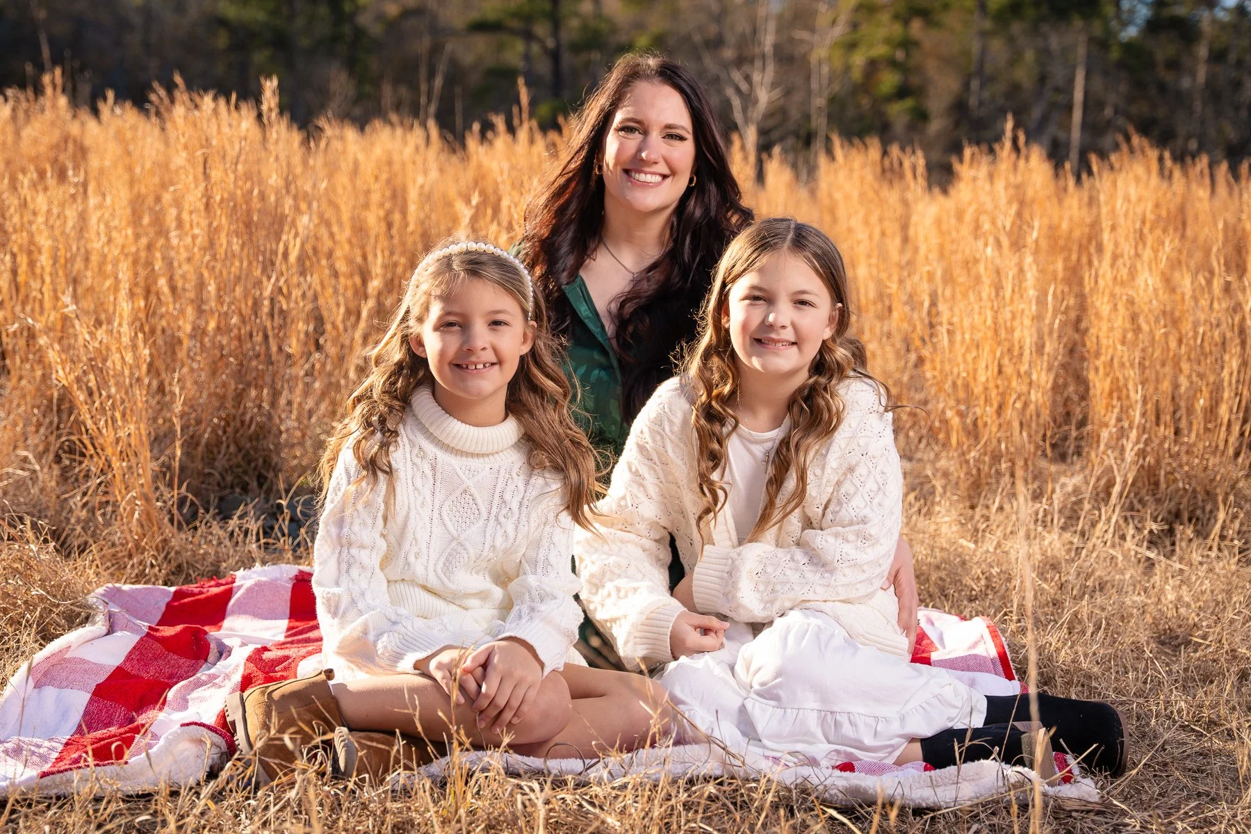 A woman and two young girls sitting on a red and white checkered blanket in a field of tall, golden grass during autumn. The woman is smiling and has long dark hair, wearing a green shirt. The girls are wearing white sweaters and skirts, with long wa