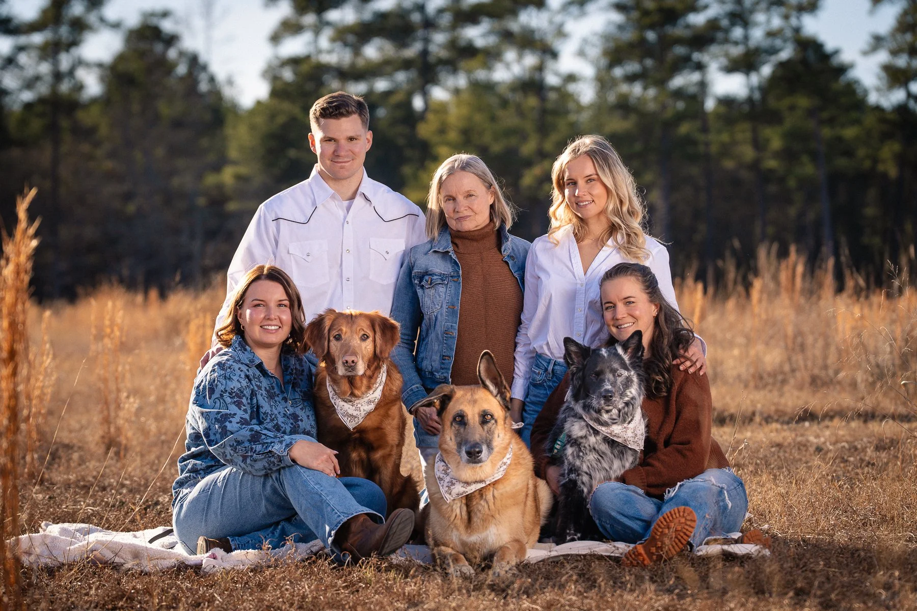 A family of six people and three dogs outdoors in a field with trees in the background, sitting on a blanket and smiling at the camera.
