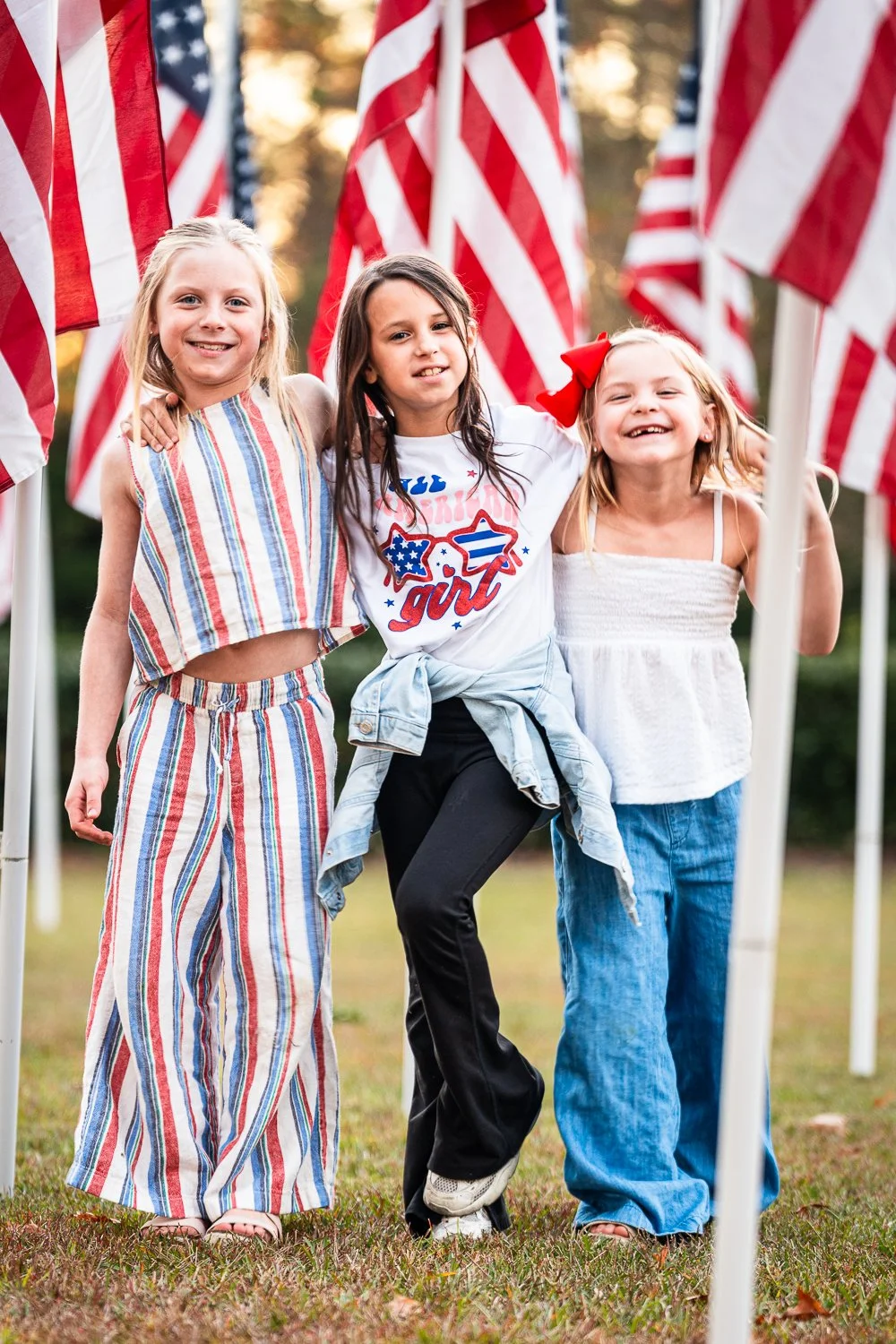 Three young girls smiling and standing together outdoors among American flags.