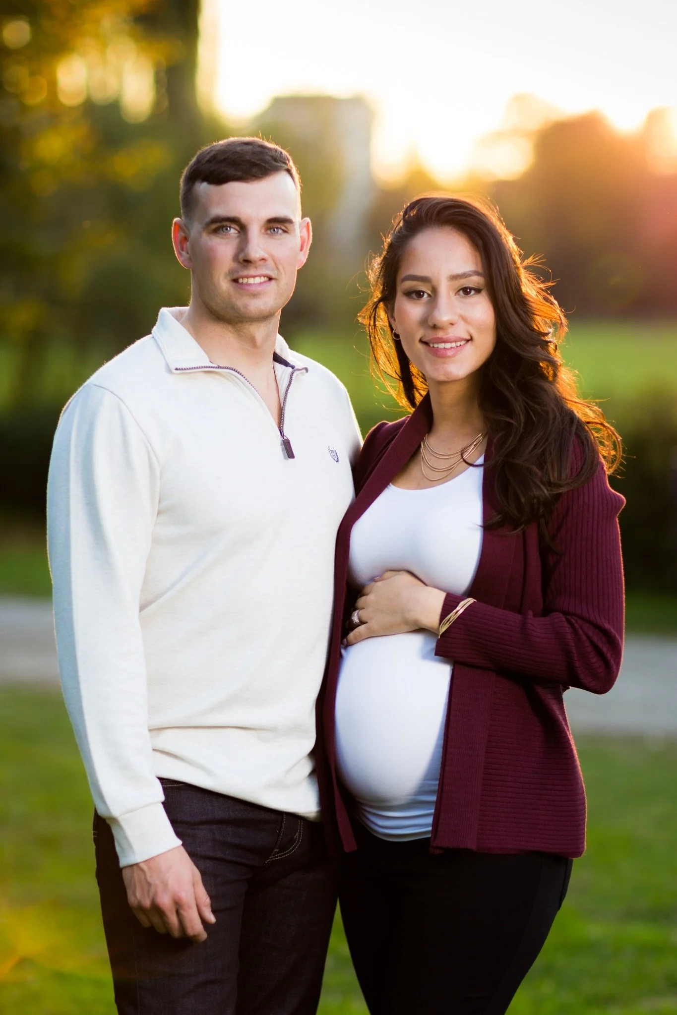 A pregnant woman and a man standing outdoors in a park during sunset, smiling and posing for a photo.