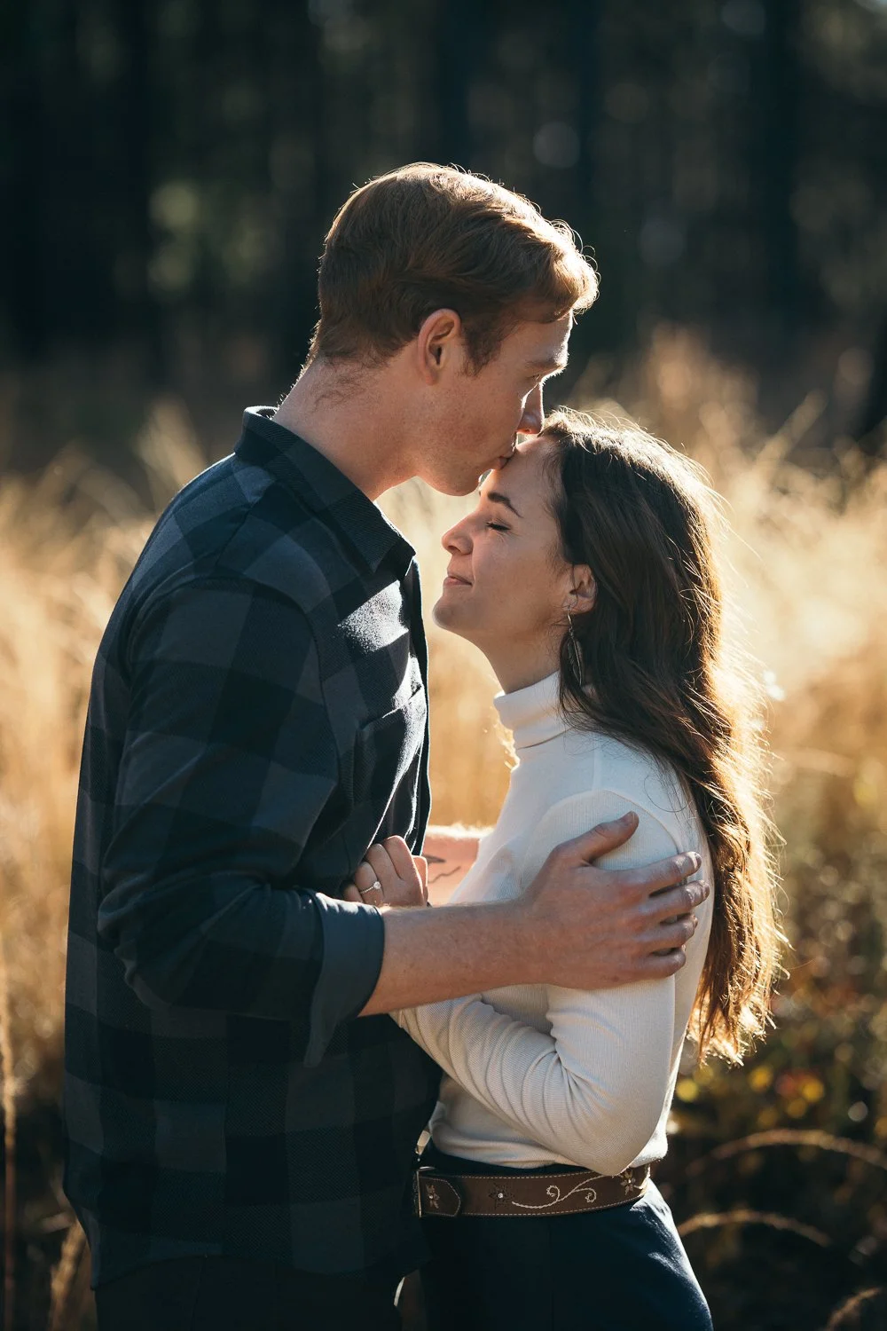 A young man kissing a young woman on the forehead in an outdoor setting during daylight, with sunlit grass and blurred trees in the background.