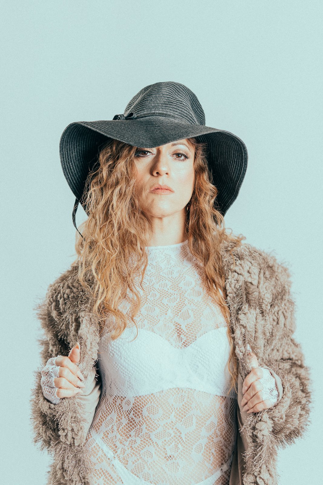 A woman with wavy red hair wearing a large black hat, a lace white top, and a fluffy coat, holding the coat's lapels, against a light background.