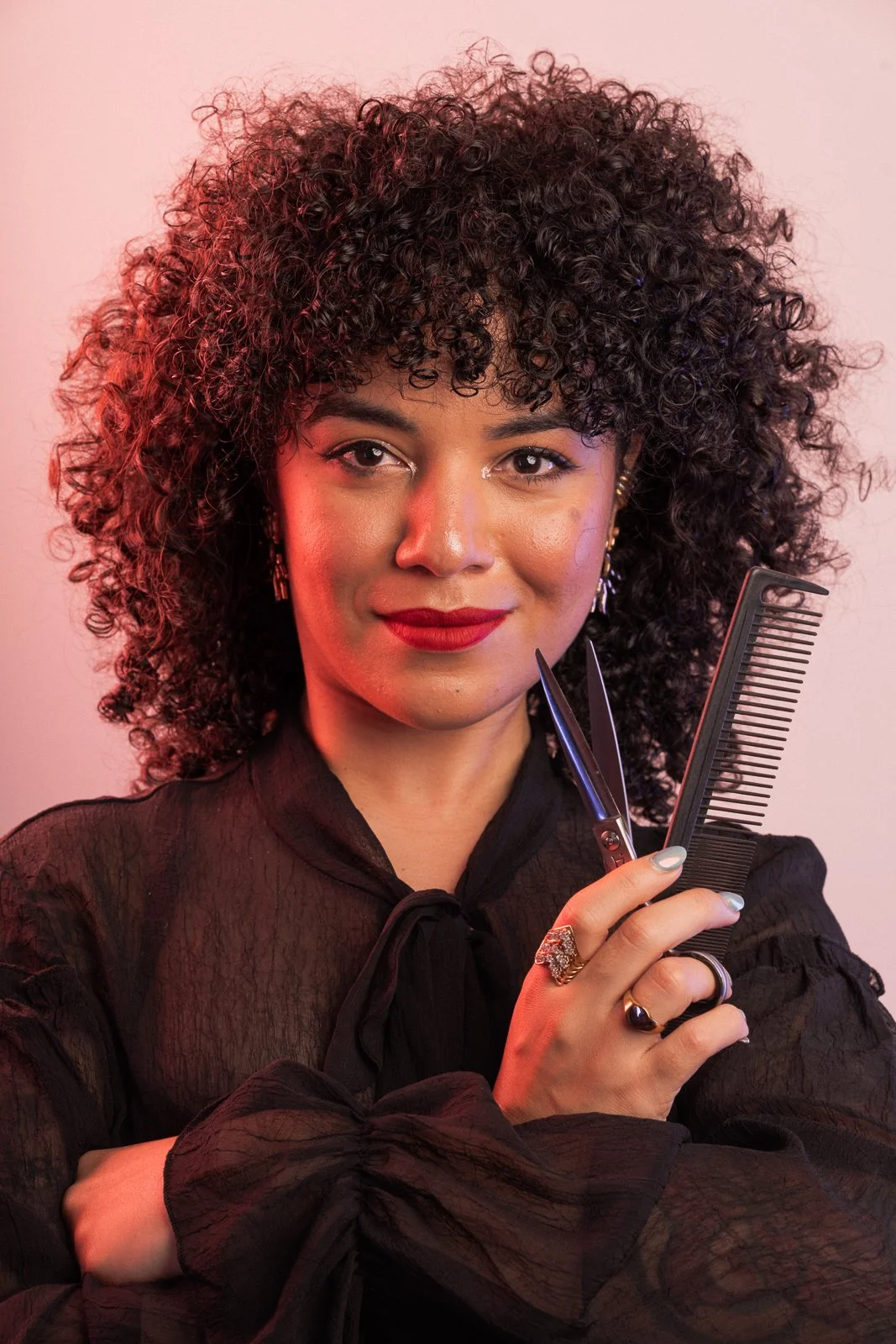 A woman with curly hair, red lipstick, and earrings, holding scissors and a comb, dressed in a black blouse, posing confidently.