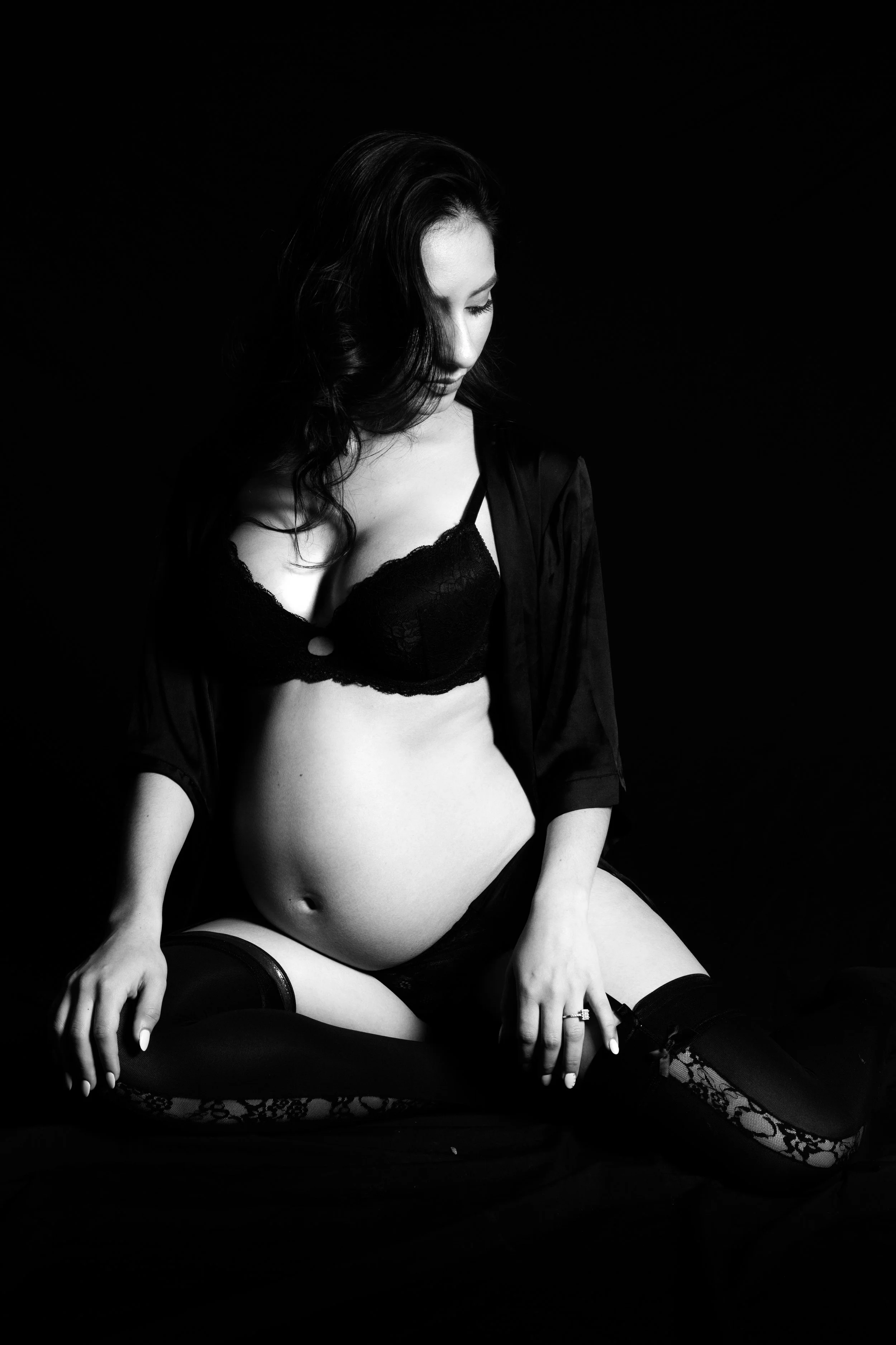 A woman dressed in black lingerie and stockings, sitting on a black surface with a dark background, looking down.