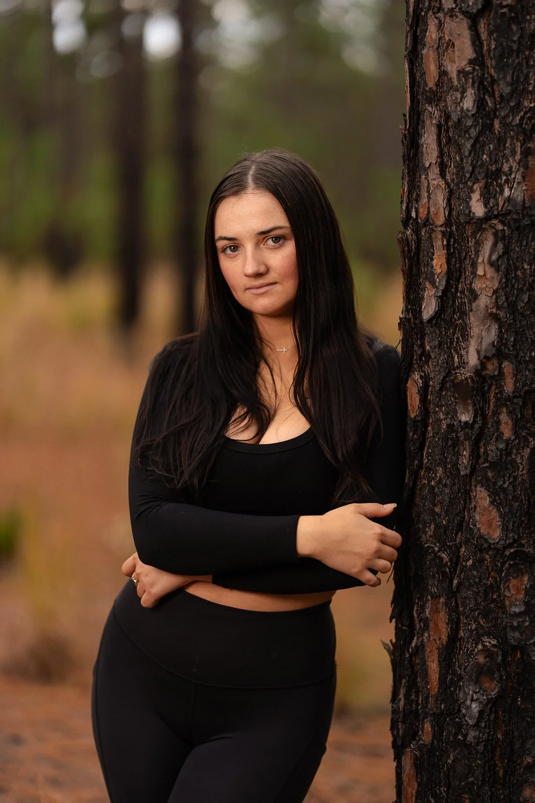 A young woman with long dark hair standing outdoors in a forest, leaning against a tree and looking at the camera.