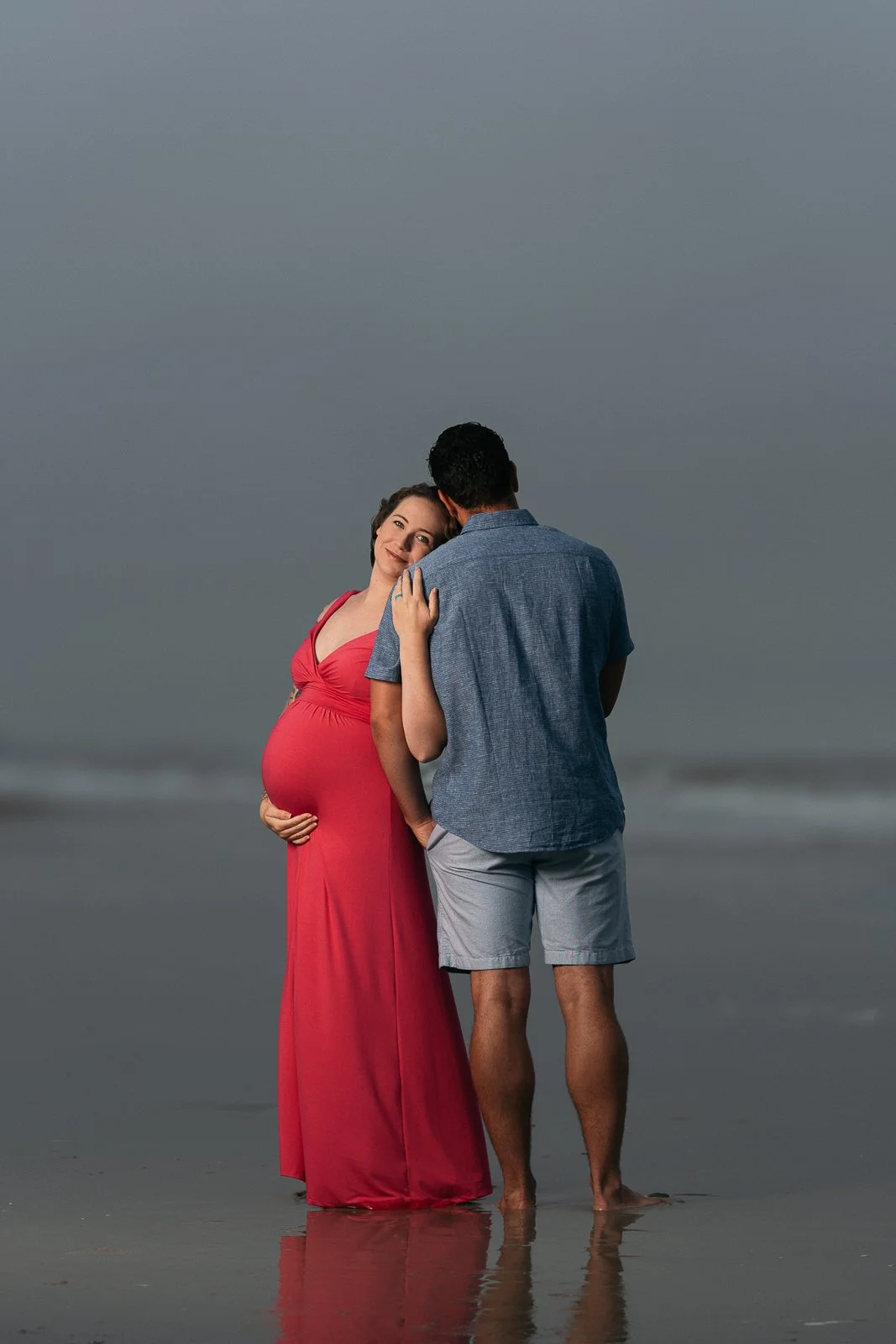 A pregnant woman in a red dress standing on the beach with a man in casual clothes, both with their feet in the water, during overcast weather.